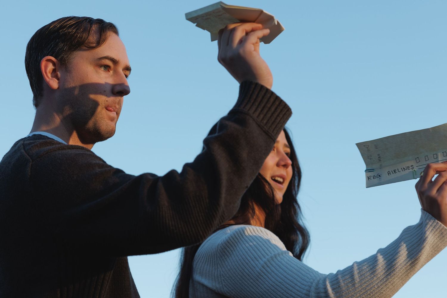 A man and woman laugh while throwing paper airplanes into a bright blue sky together.
