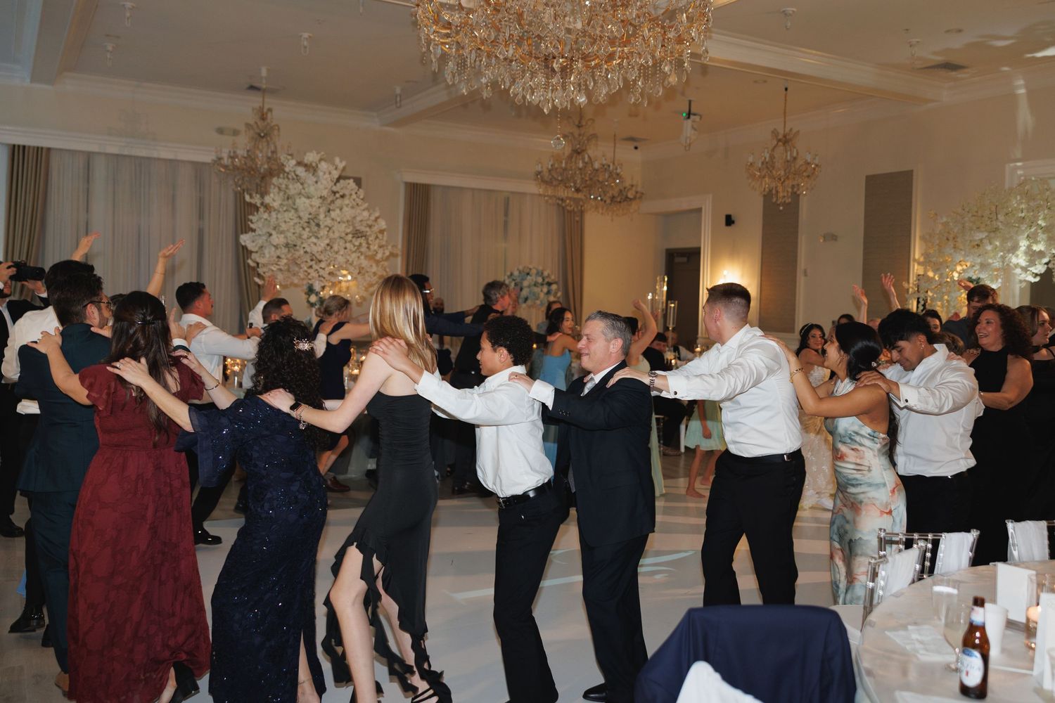 Guests form a large circle dancing together in elegant ballroom with chandeliers at wedding reception.