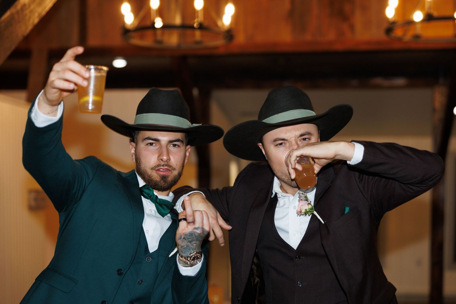 Two groomsmen in green and black suits with cowboy hats toasting drinks at wedding.