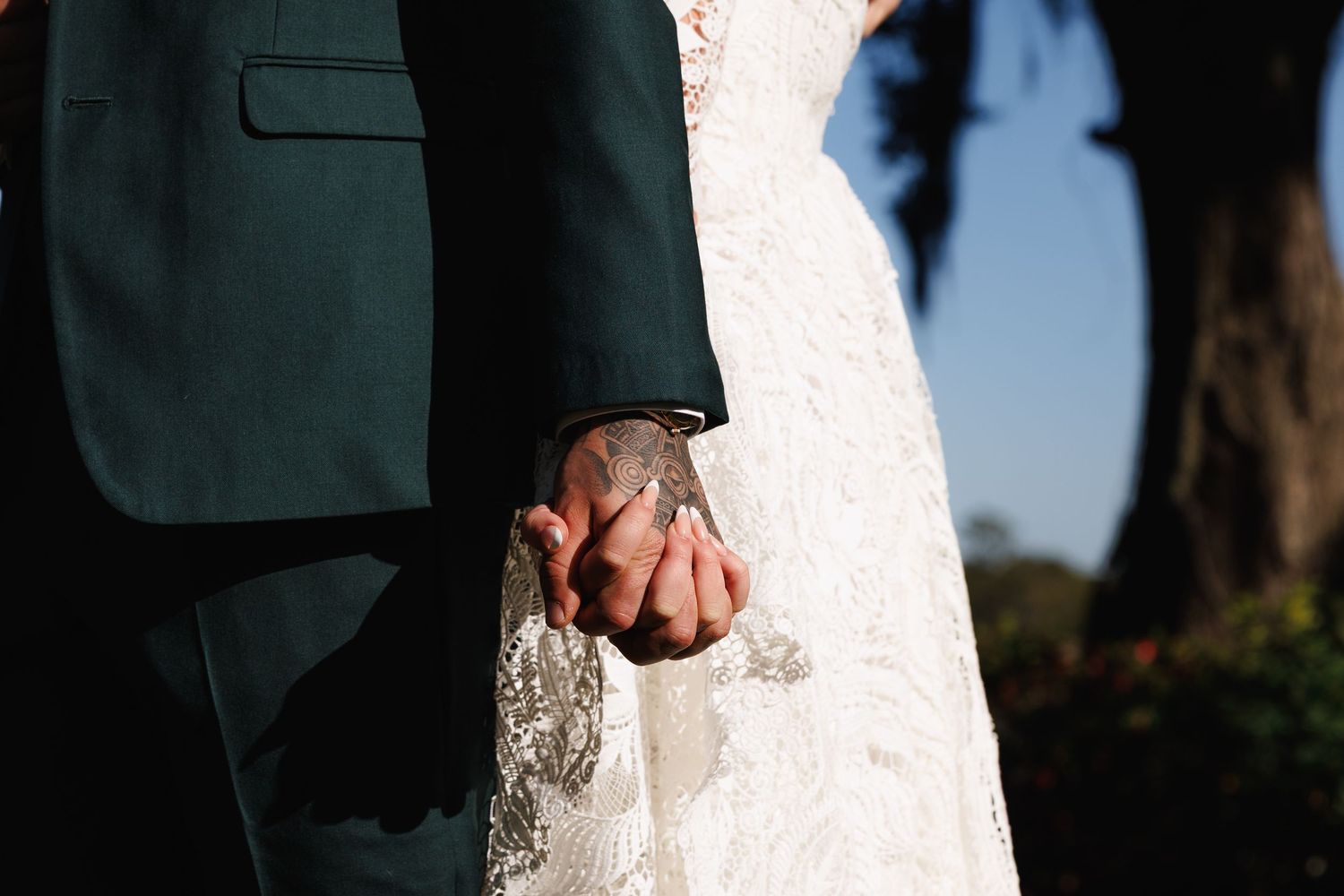 Close-up of bride and groom holding hands, showcasing lace wedding dress and dark suit outdoors.