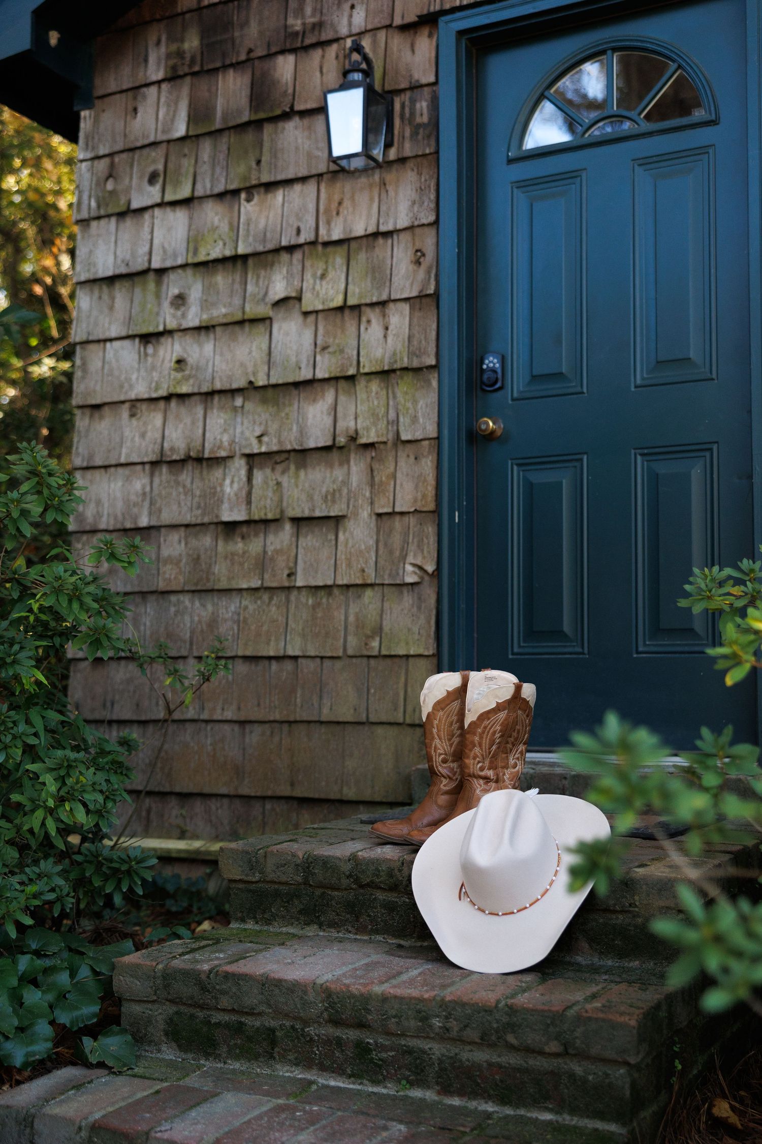 Cowboy boots and white hat on brick steps outside a rustic cedar shake home with a dark teal front door.