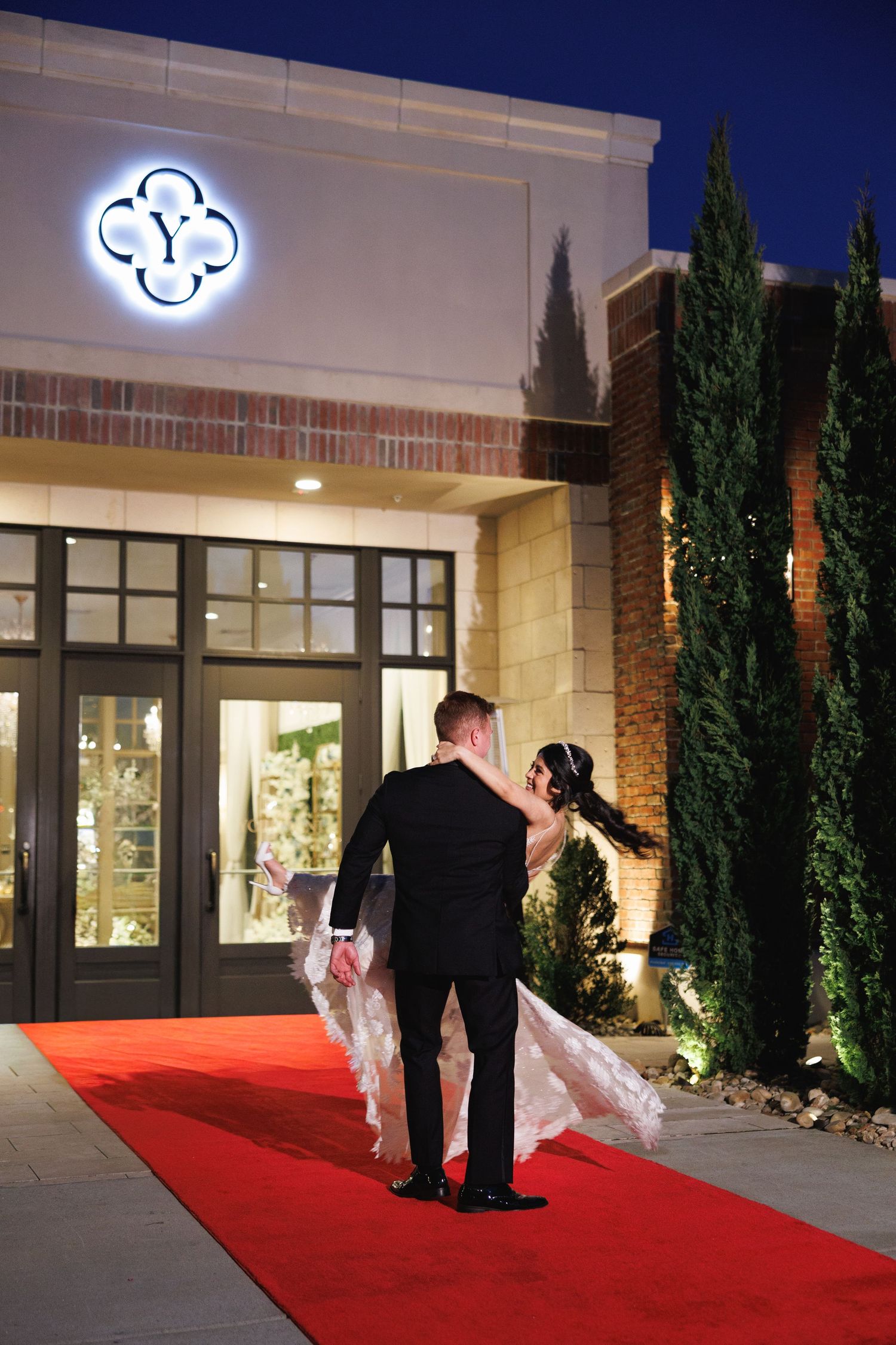 Bride and groom walk hand-in-hand on red carpet outside illuminated Yorkmont in Charlotte, North Carolina, at night.