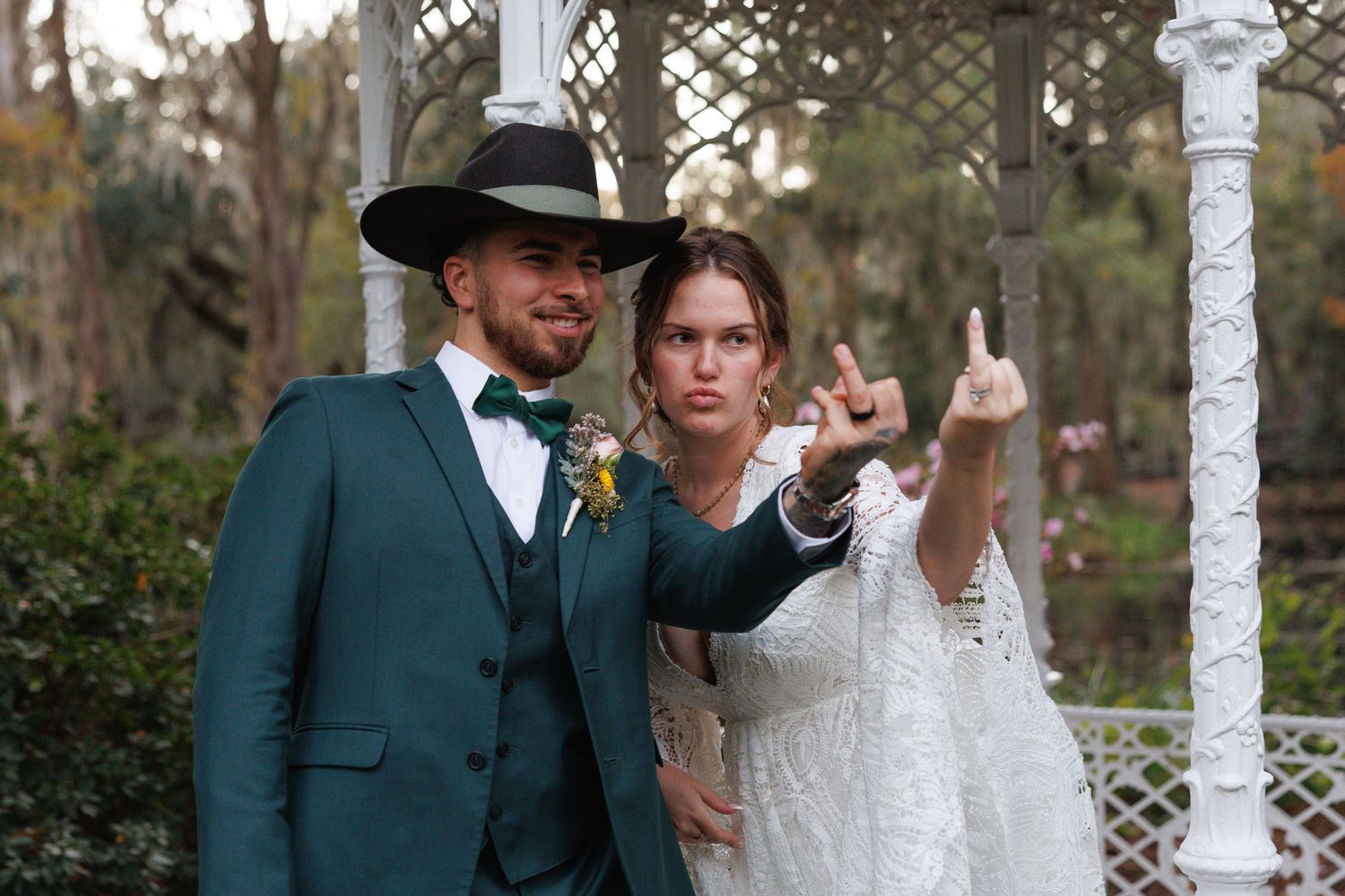 Bride and groom pose playfully in a white gazebo, groom wearing a teal suit and black cowboy hat.