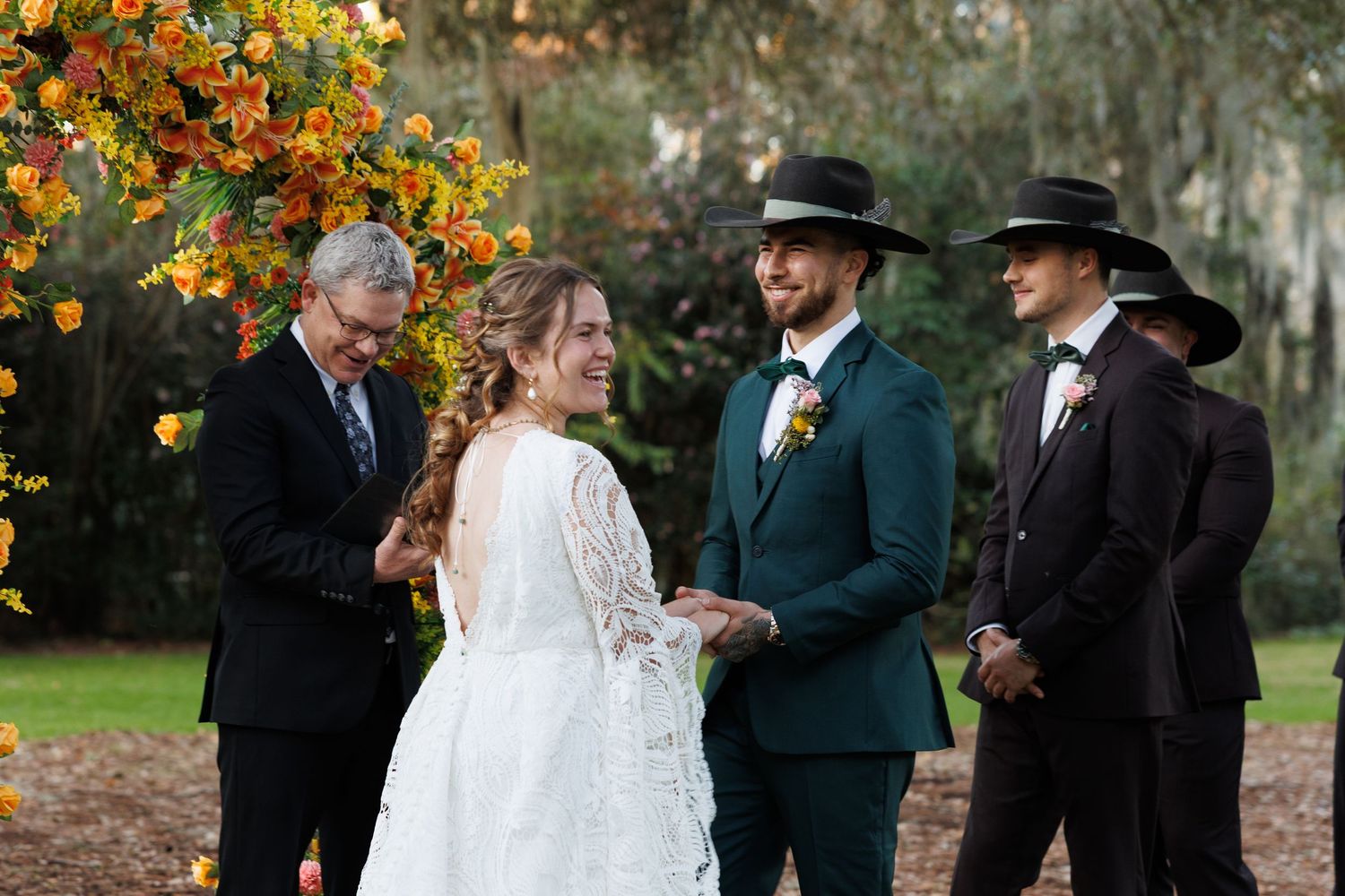 A bride and groom exchange vows outdoors under a floral arch, with groomsmen in stylish hats standing nearby.