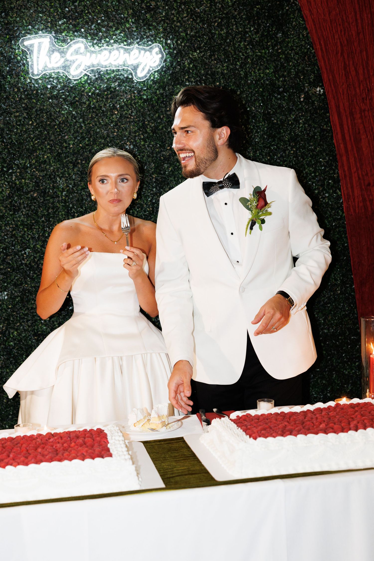 Bride and groom cut white wedding cake with raspberry decor under neon sign backdrop.