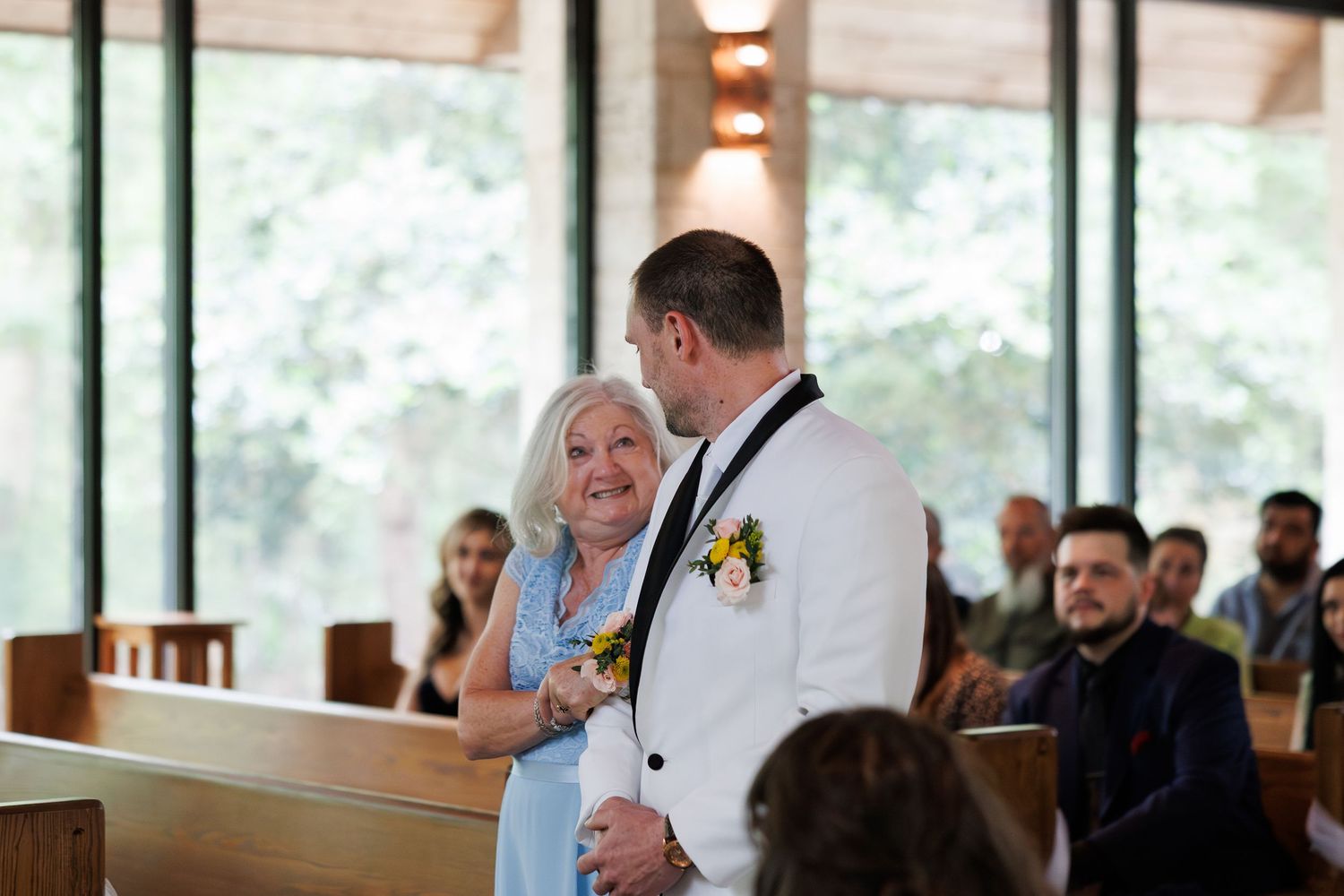 Groom in white tuxedo shares a tender moment with elderly woman in blue dress during wedding ceremony.