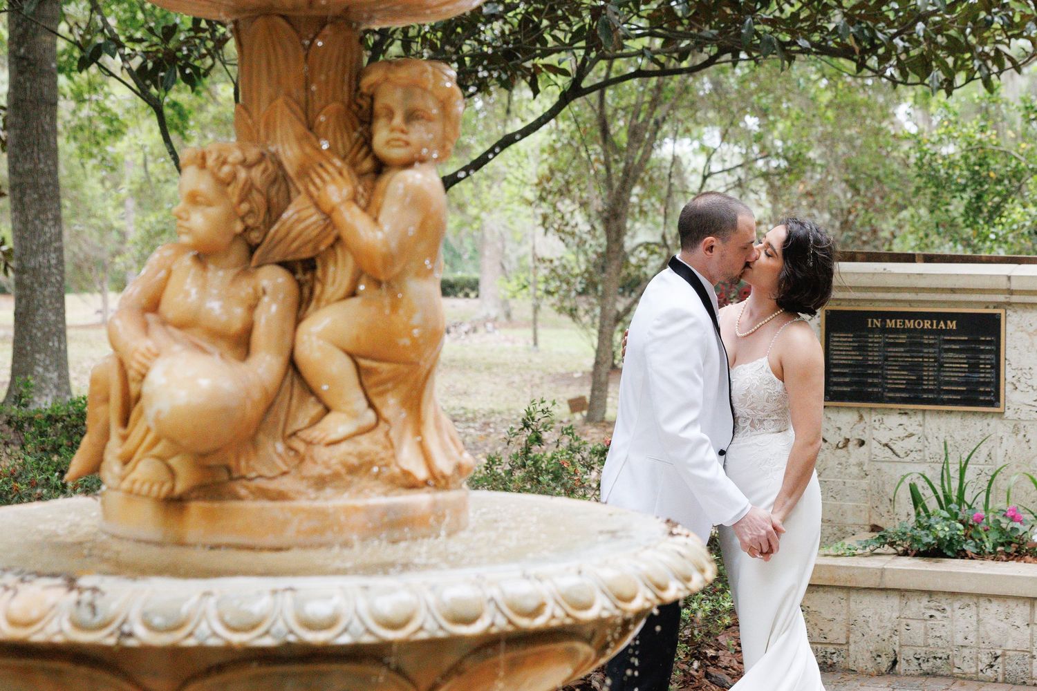 Bride and groom kissing beside ornate cherub fountain in lush garden wedding venue.