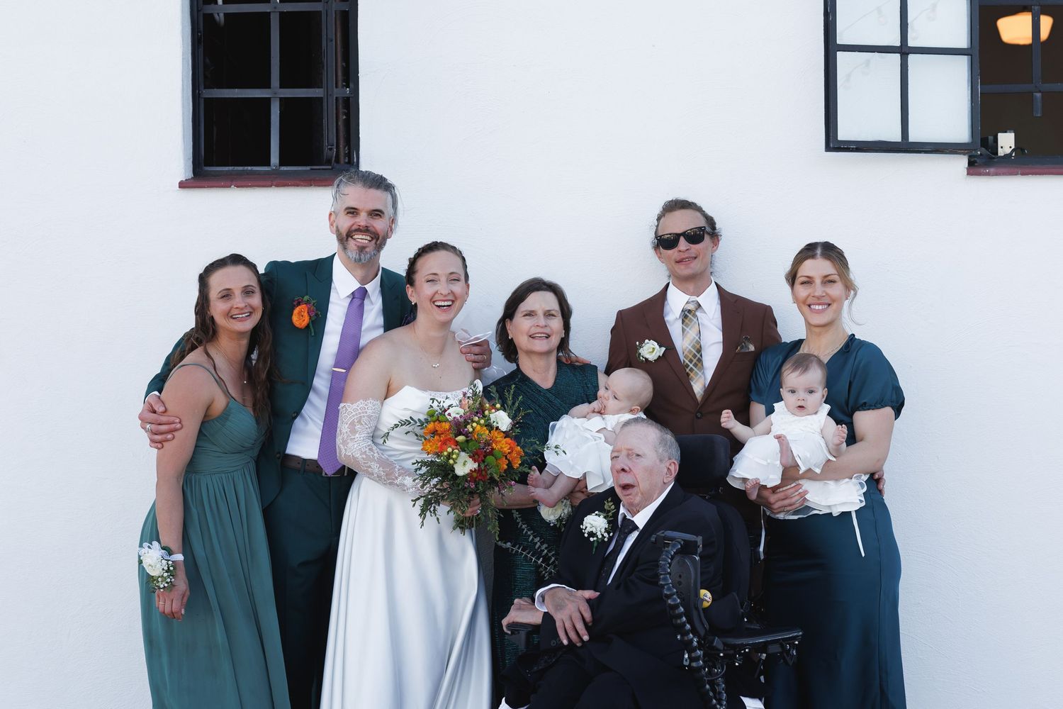 Wedding family portrait with bride in white gown, groom, bridesmaids, and guests posing outdoors against white wall.