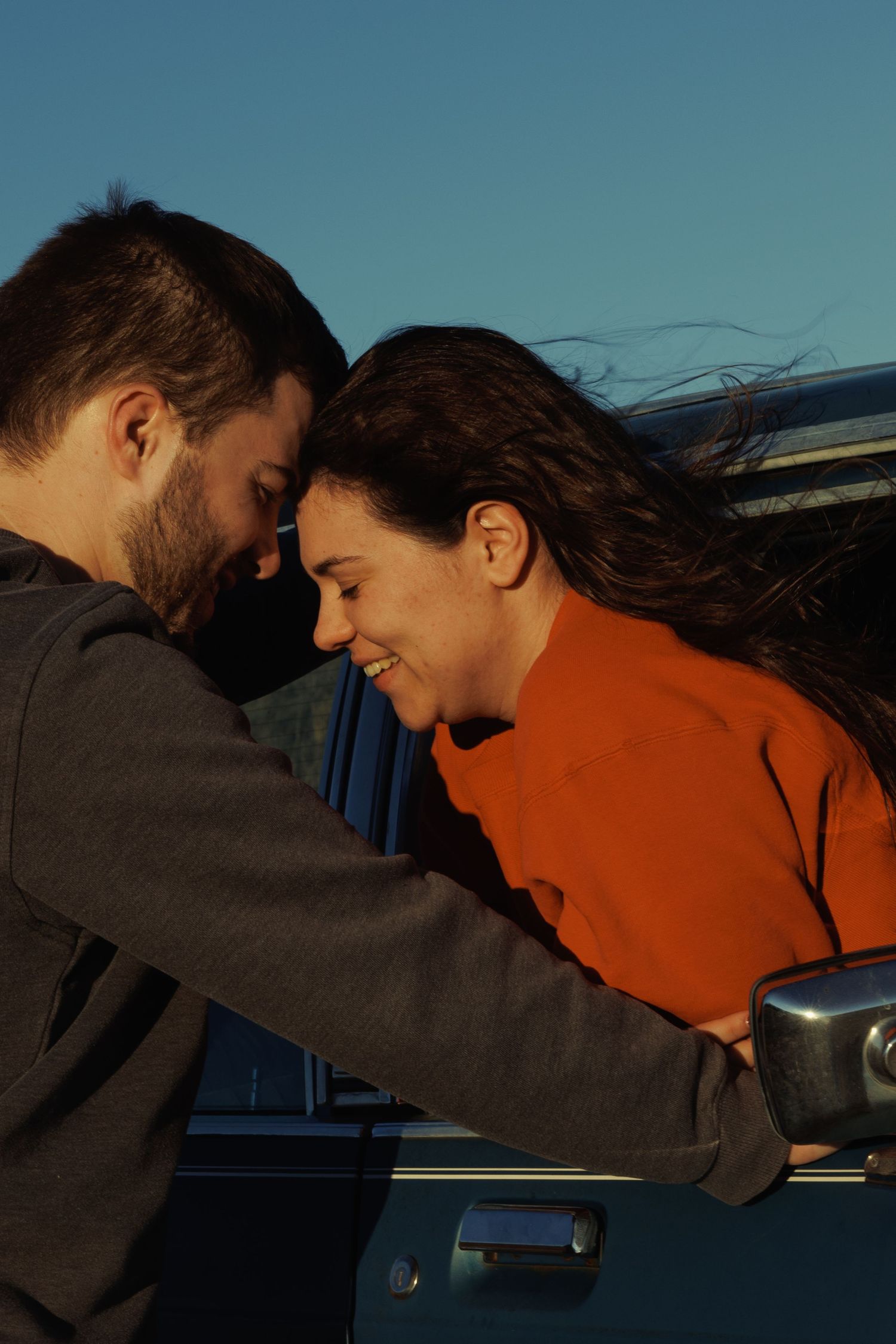 Couple sharing a romantic moment leaning foreheads together beside a vintage blue van at sunset.