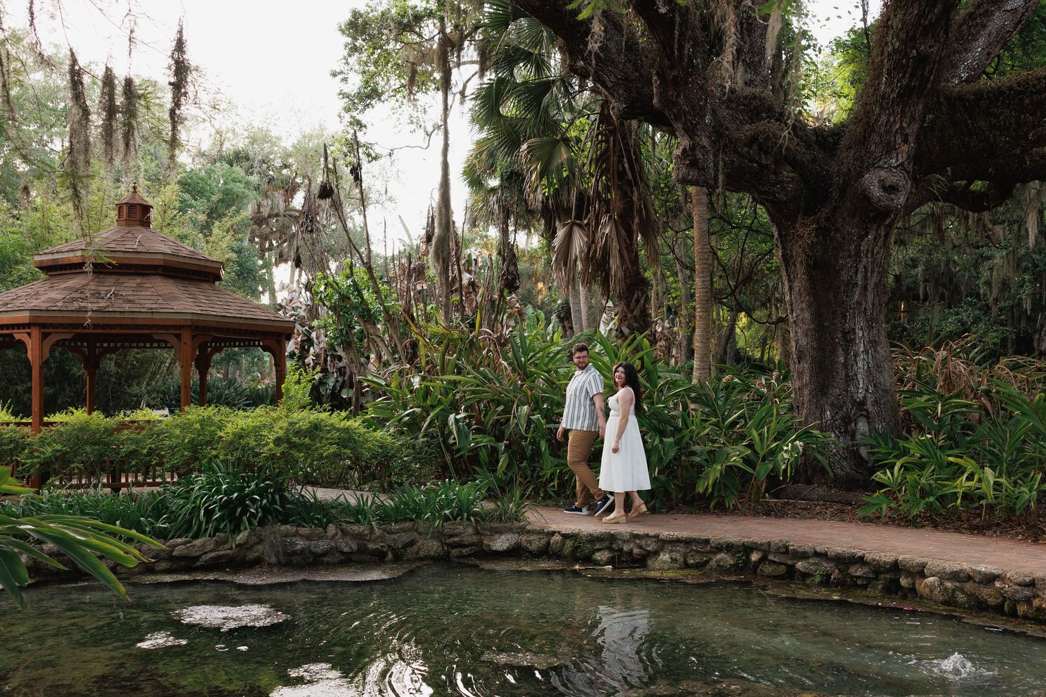 Couple standing by a serene pond in a lush tropical garden with a gazebo and large ancient tree.