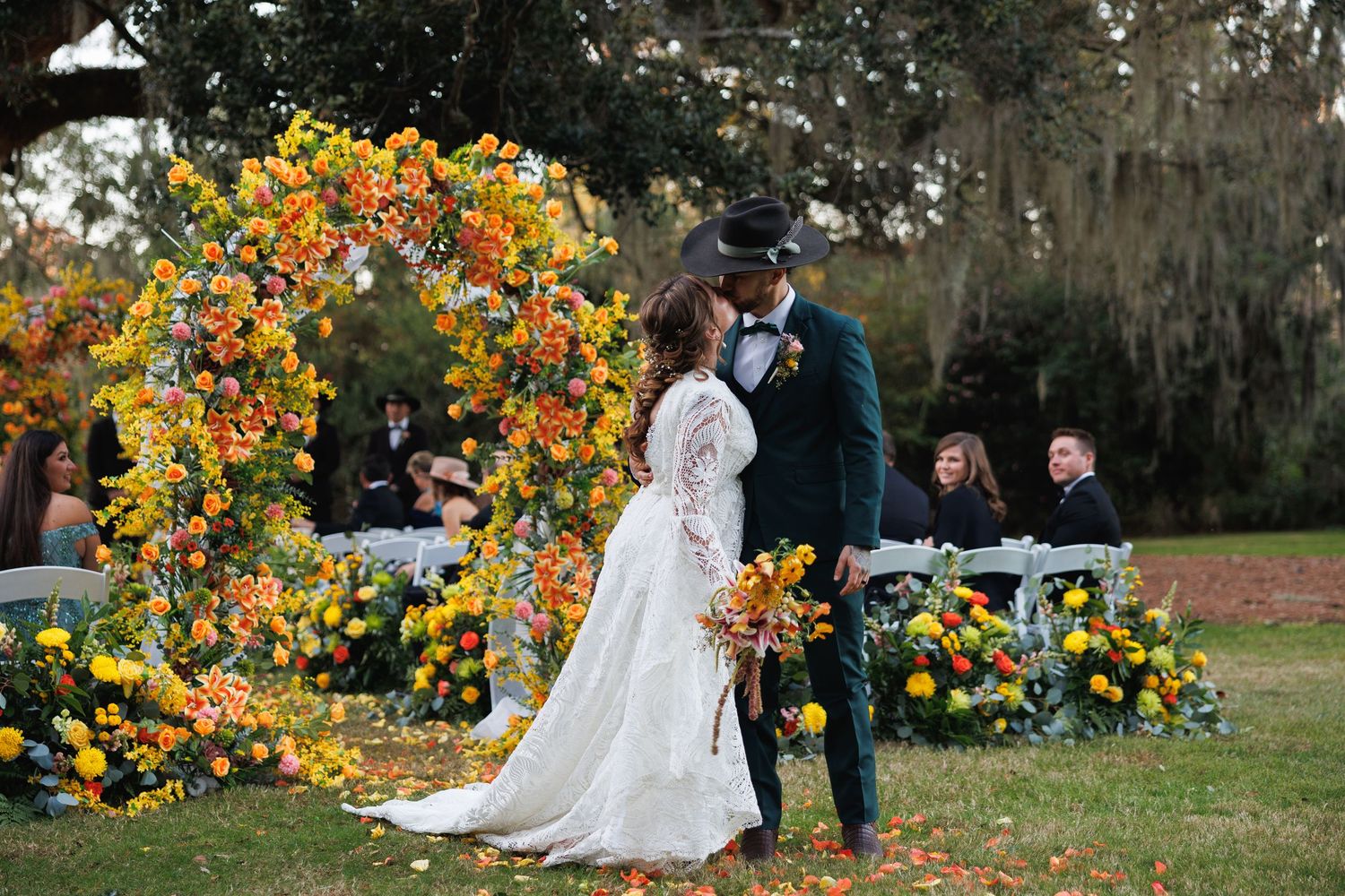 Newlyweds share a first kiss under a vibrant autumn floral arch at an elegant outdoor wedding ceremony.