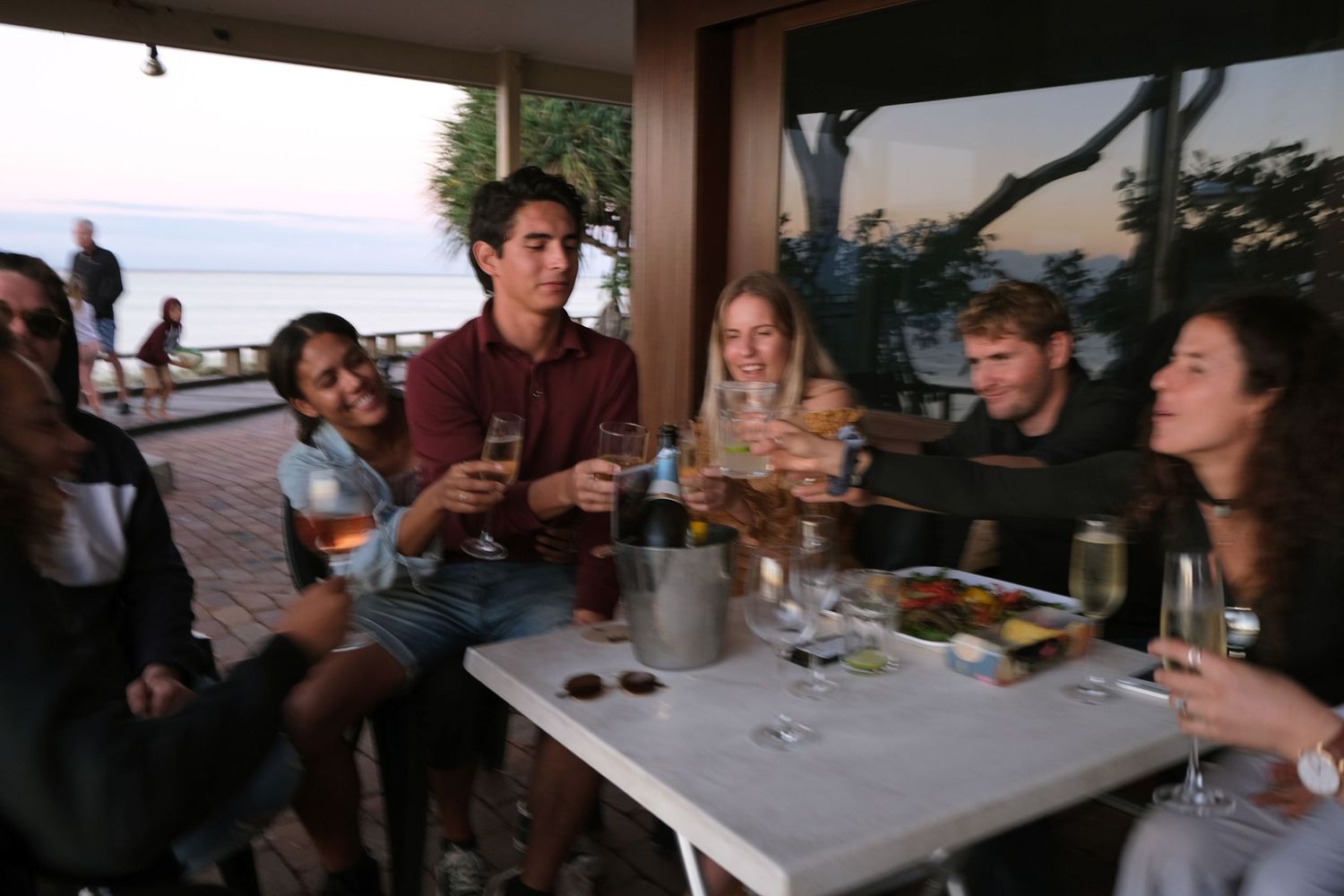 Group of young friends toasting with wine and champagne at an outdoor restaurant with ocean views at sunset.