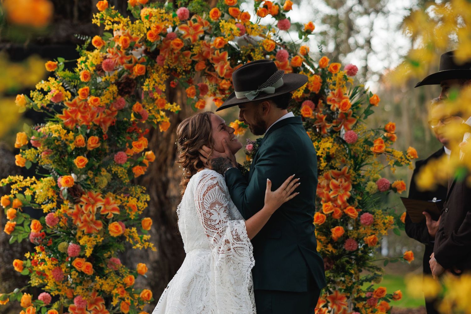 Couple sharing romantic kiss under vibrant orange and yellow floral arch at outdoor fall wedding ceremony.