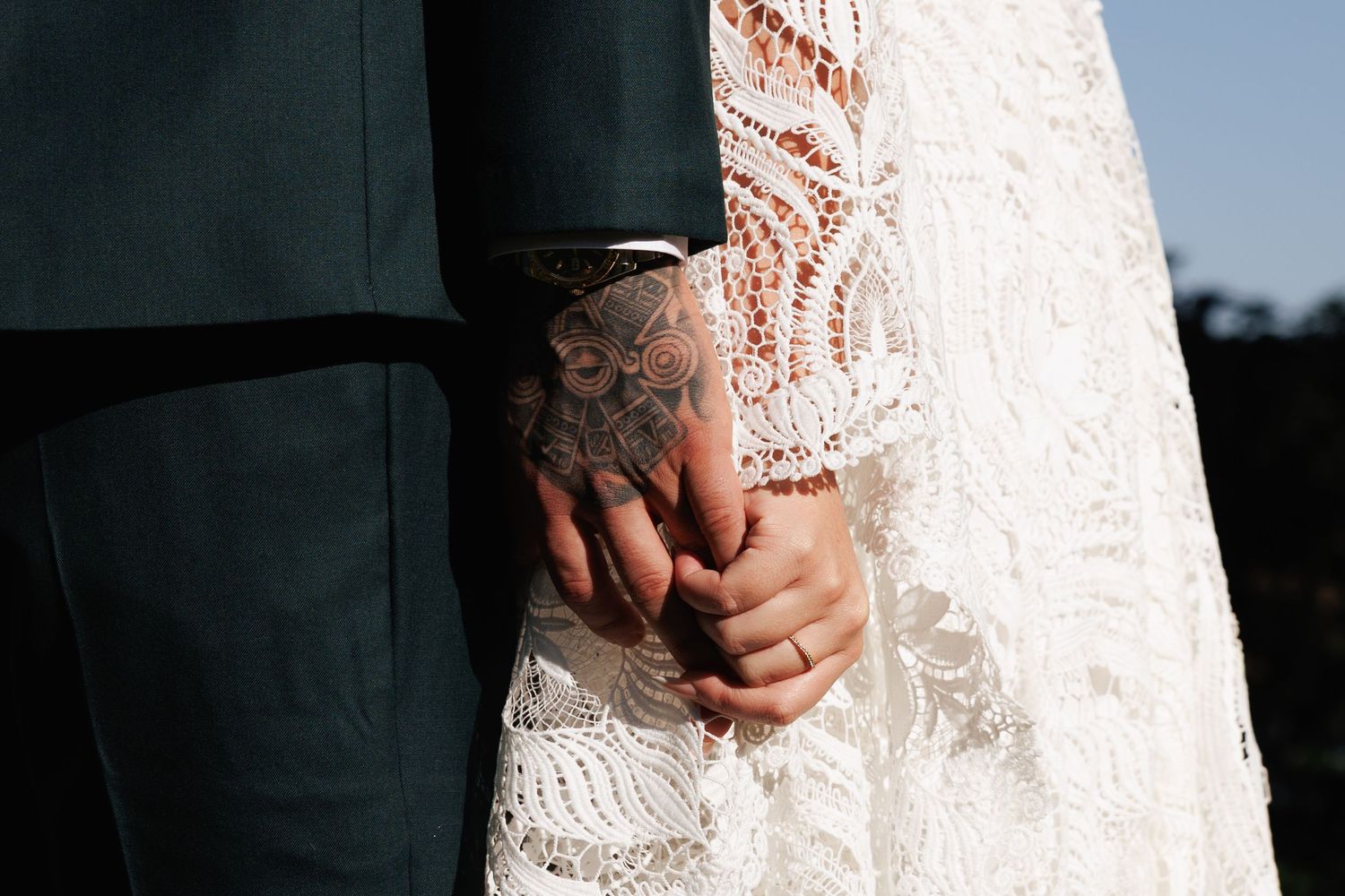 Close-up of a tattooed groom holding hands with a bride in a lace wedding dress.