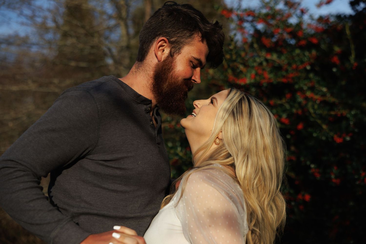 A bearded man and blonde woman share a romantic almost-kiss moment in a garden with red berries.