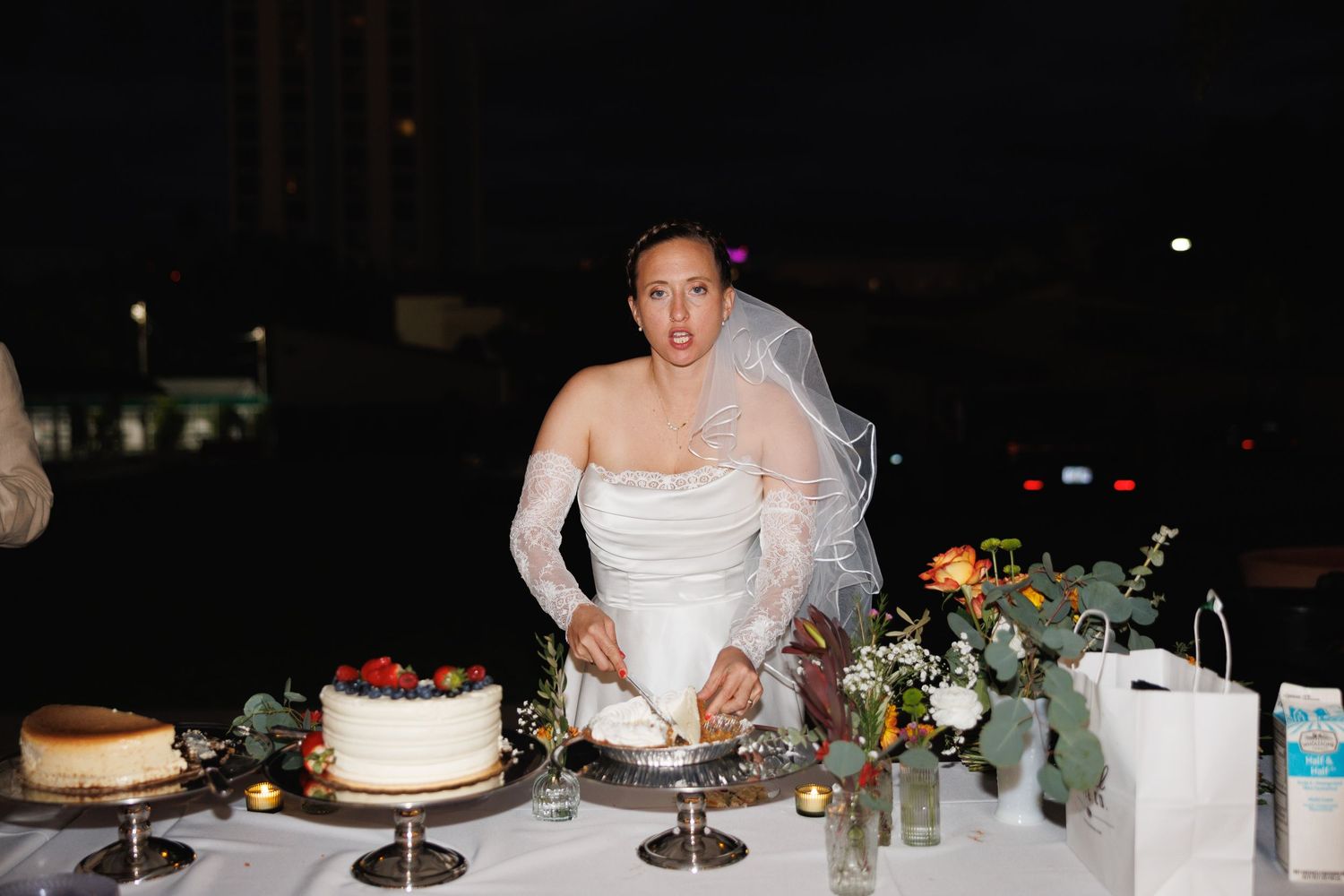 Bride in white gown and veil cuts wedding cake at night reception table with floral decorations.