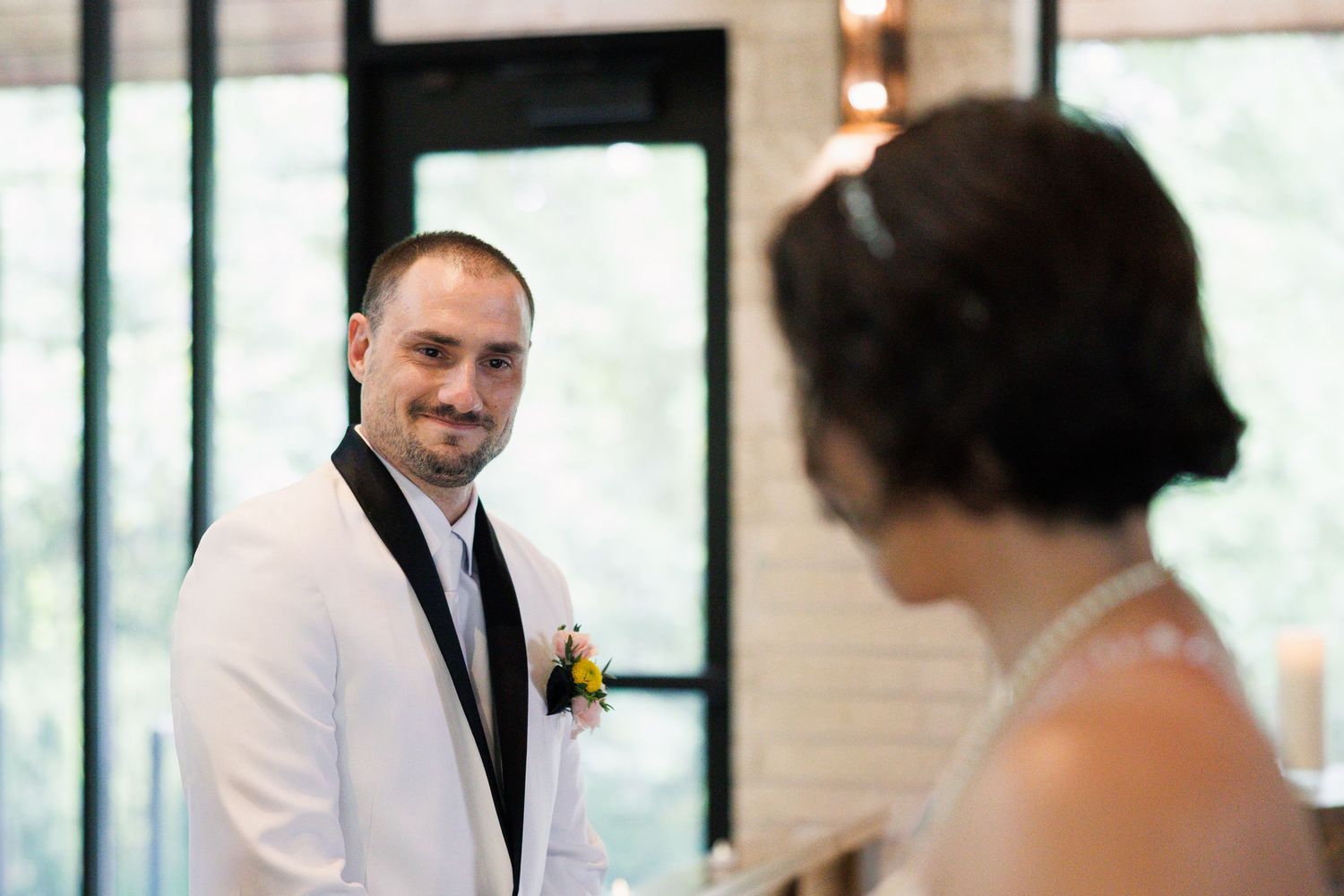 Groom in white tuxedo smiling at bride during wedding ceremony.