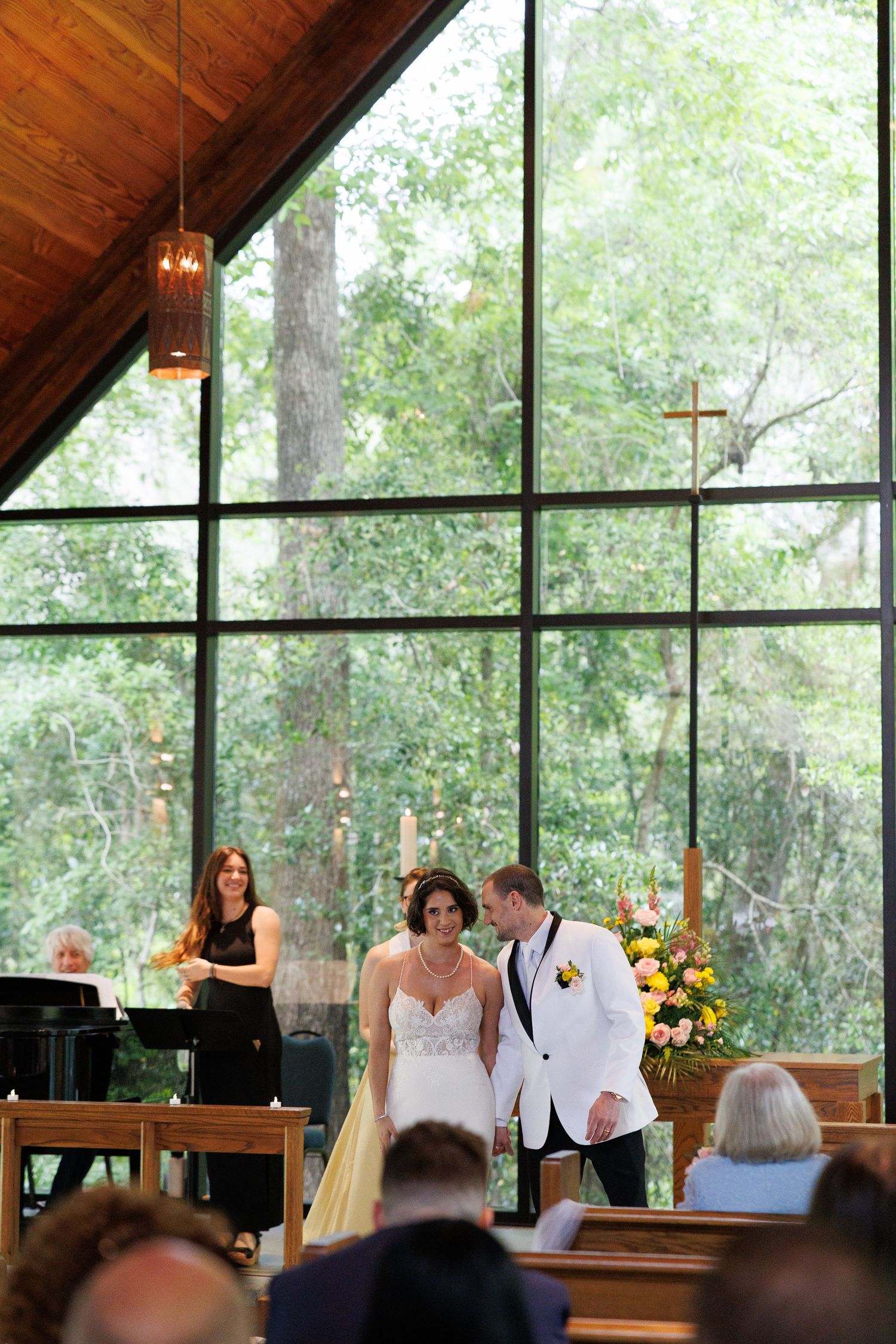 Bride and groom stand at the altar of a chapel with large windows overlooking lush green trees.