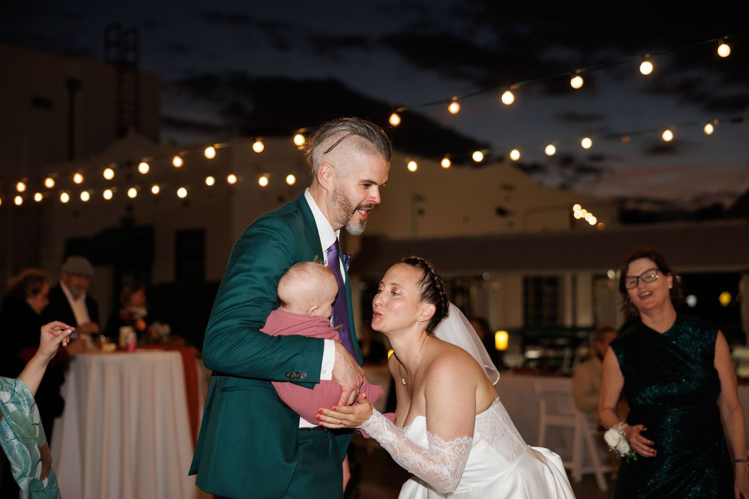 Groom in teal suit holds baby while bride in white gown smiles at outdoor evening wedding reception.