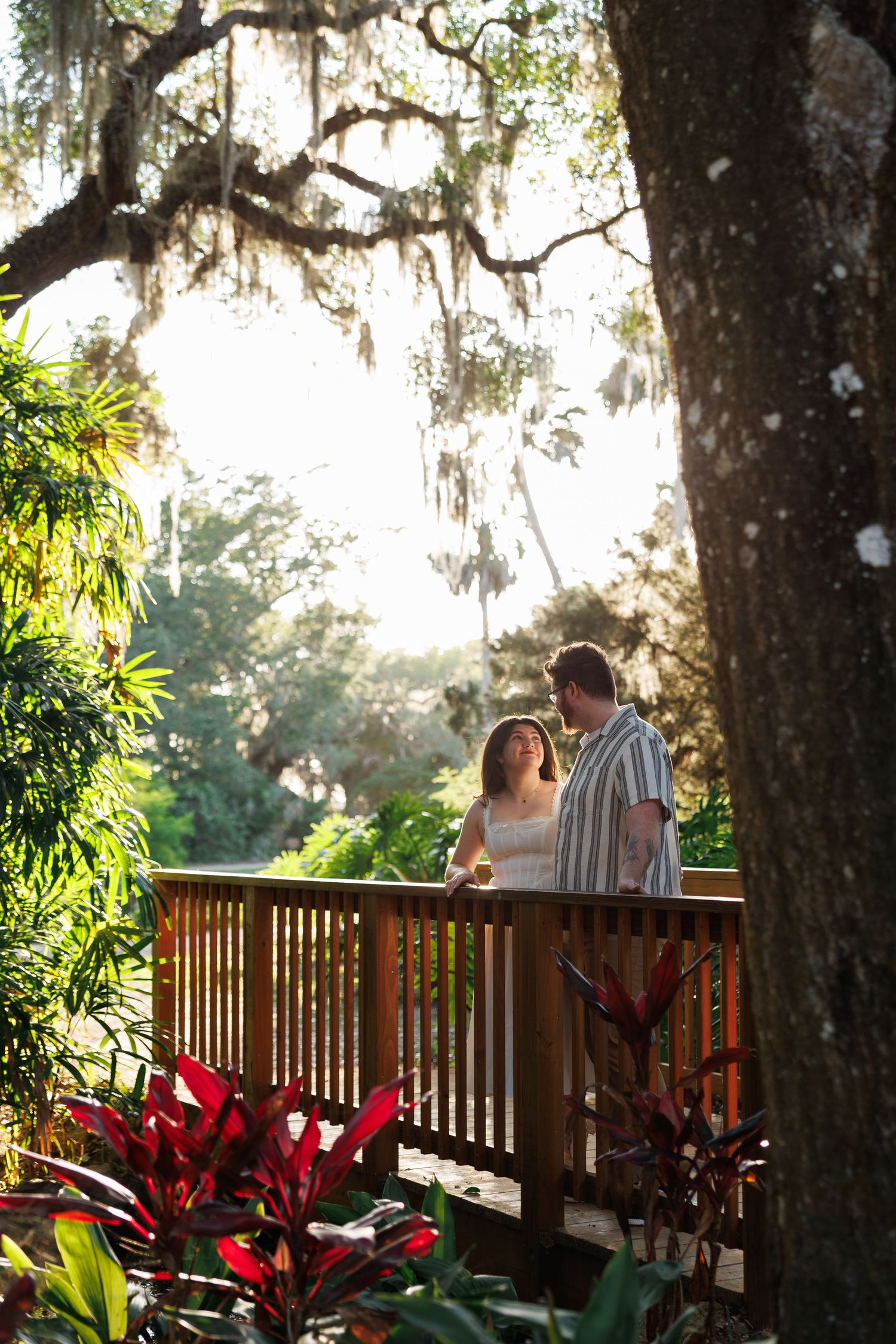 Couple standing on a wooden deck surrounded by lush tropical greenery and Spanish moss-draped oak trees.