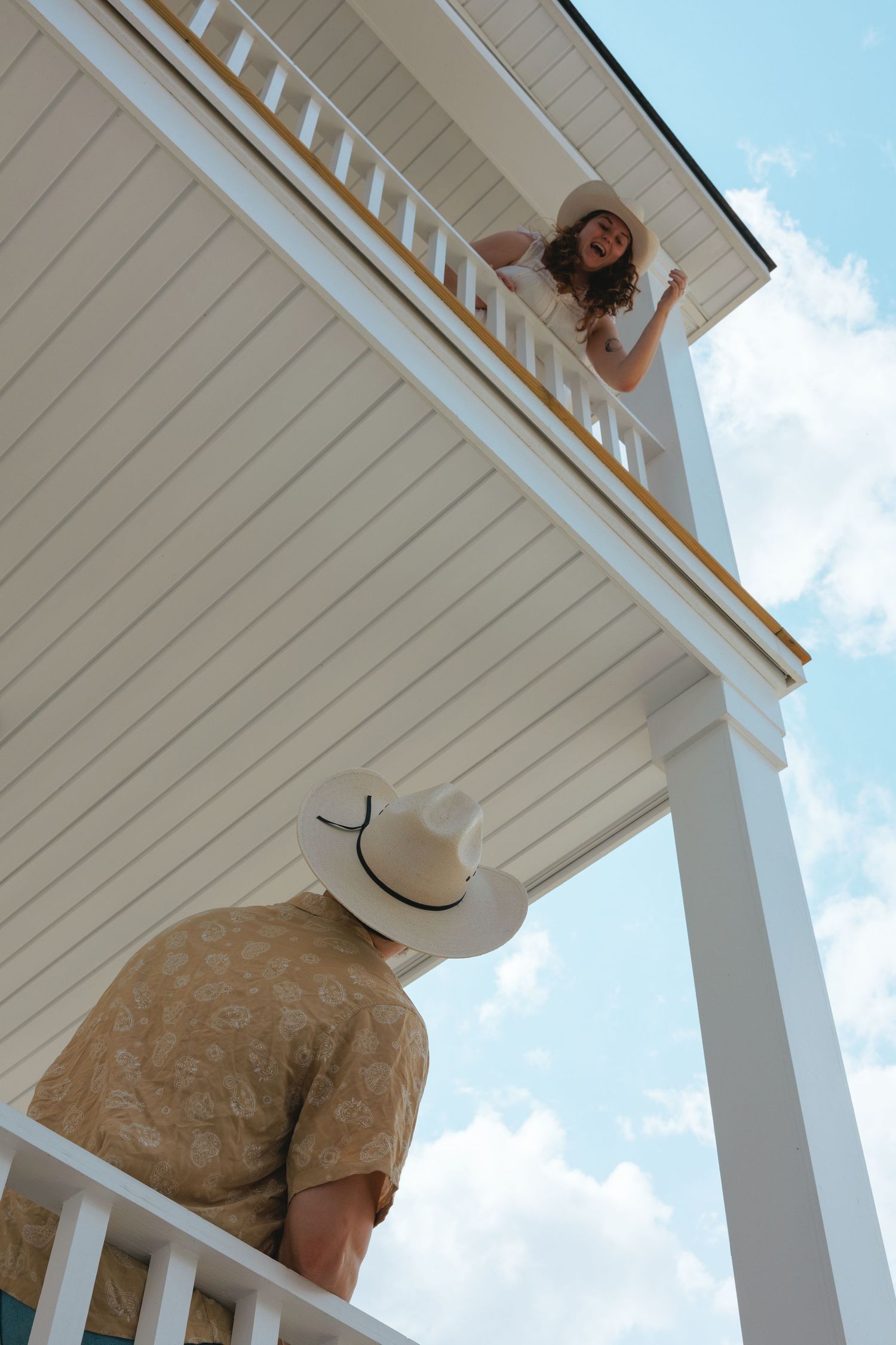 Western engagement session with fiance looking up at his fiance on second floor balcony of white house by Gainesville wedding photographer.