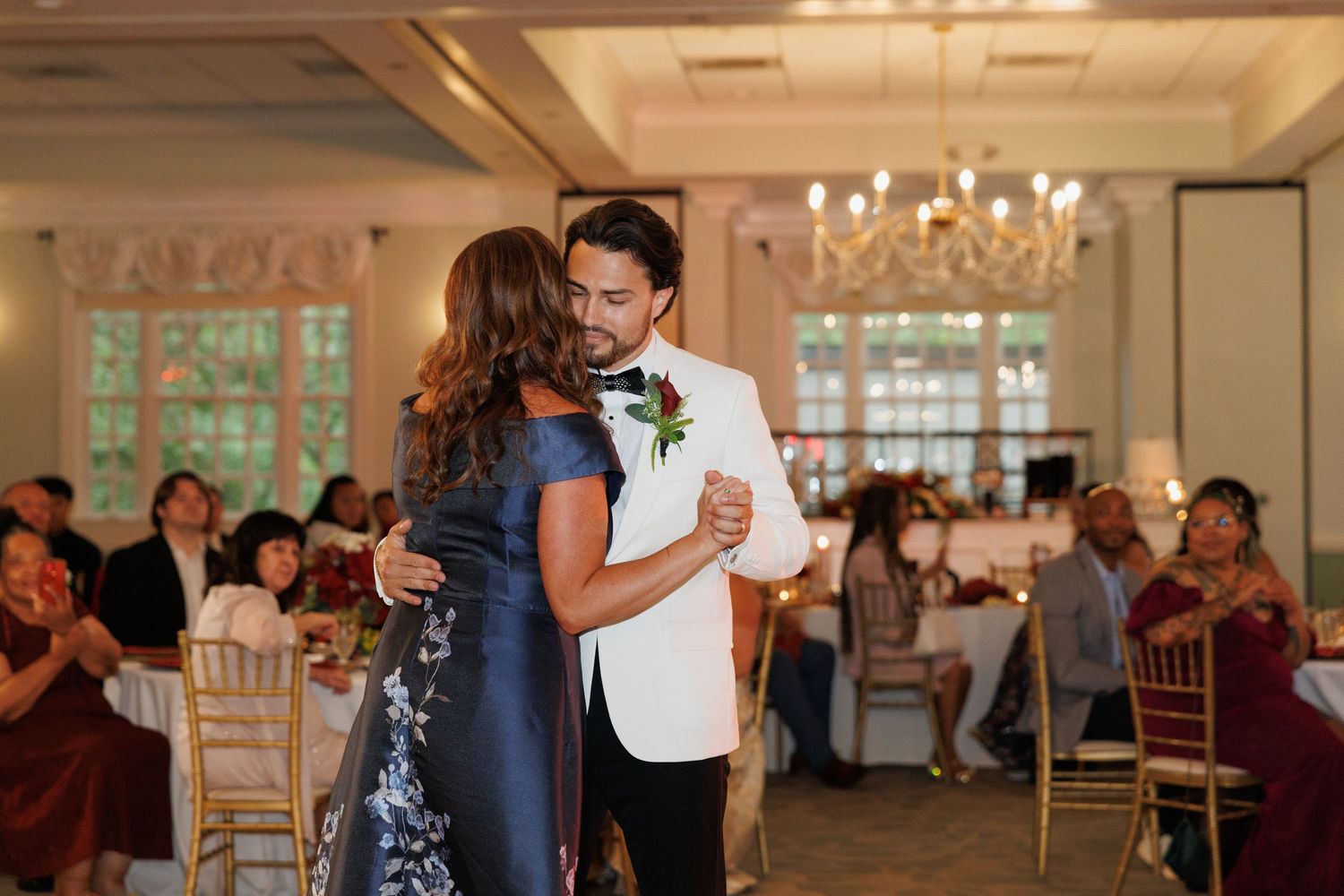 Groom in white tuxedo shares first dance with mother at elegant wedding reception with chandelier and seated guests.