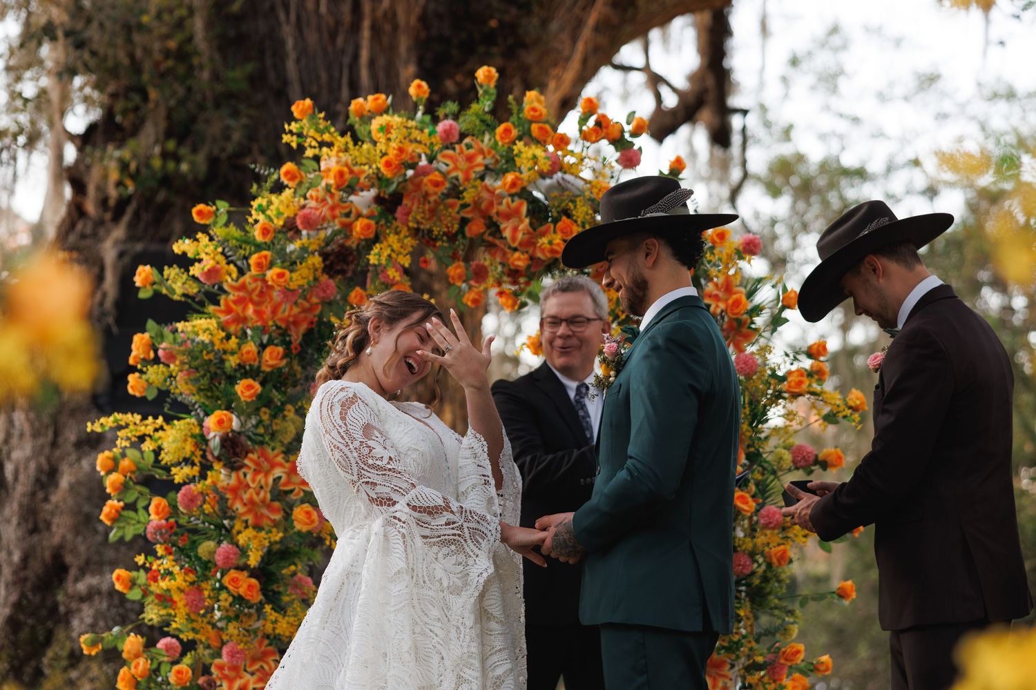 Couple exchanges vows at colorful outdoor wedding ceremony with vibrant orange and yellow floral arch backdrop.