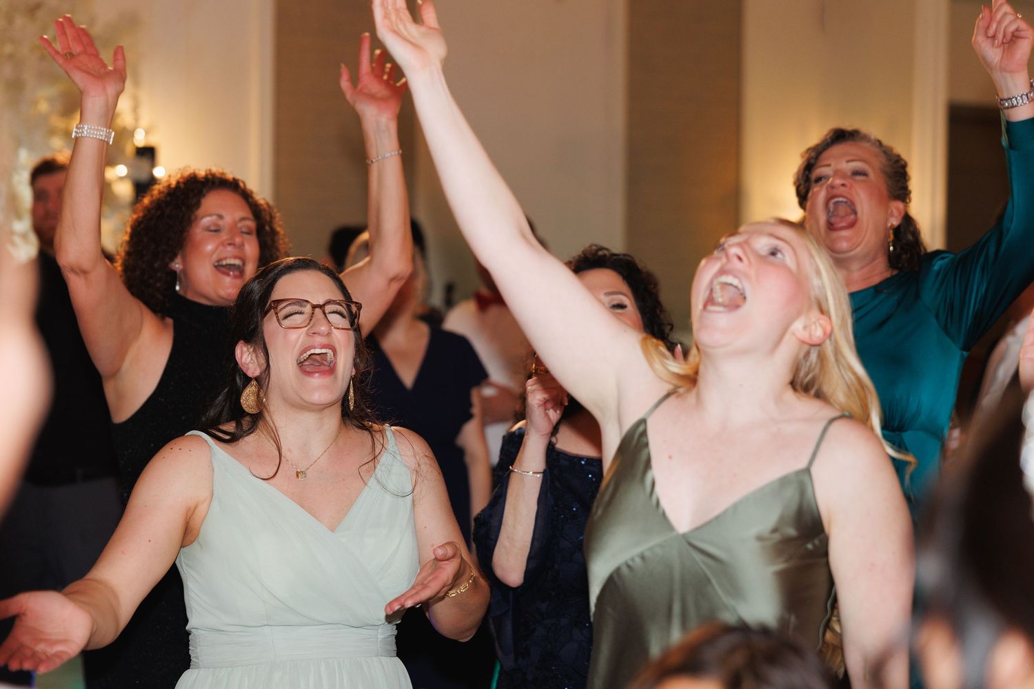 Excited wedding guests raise their arms and sing along on the reception dance floor.