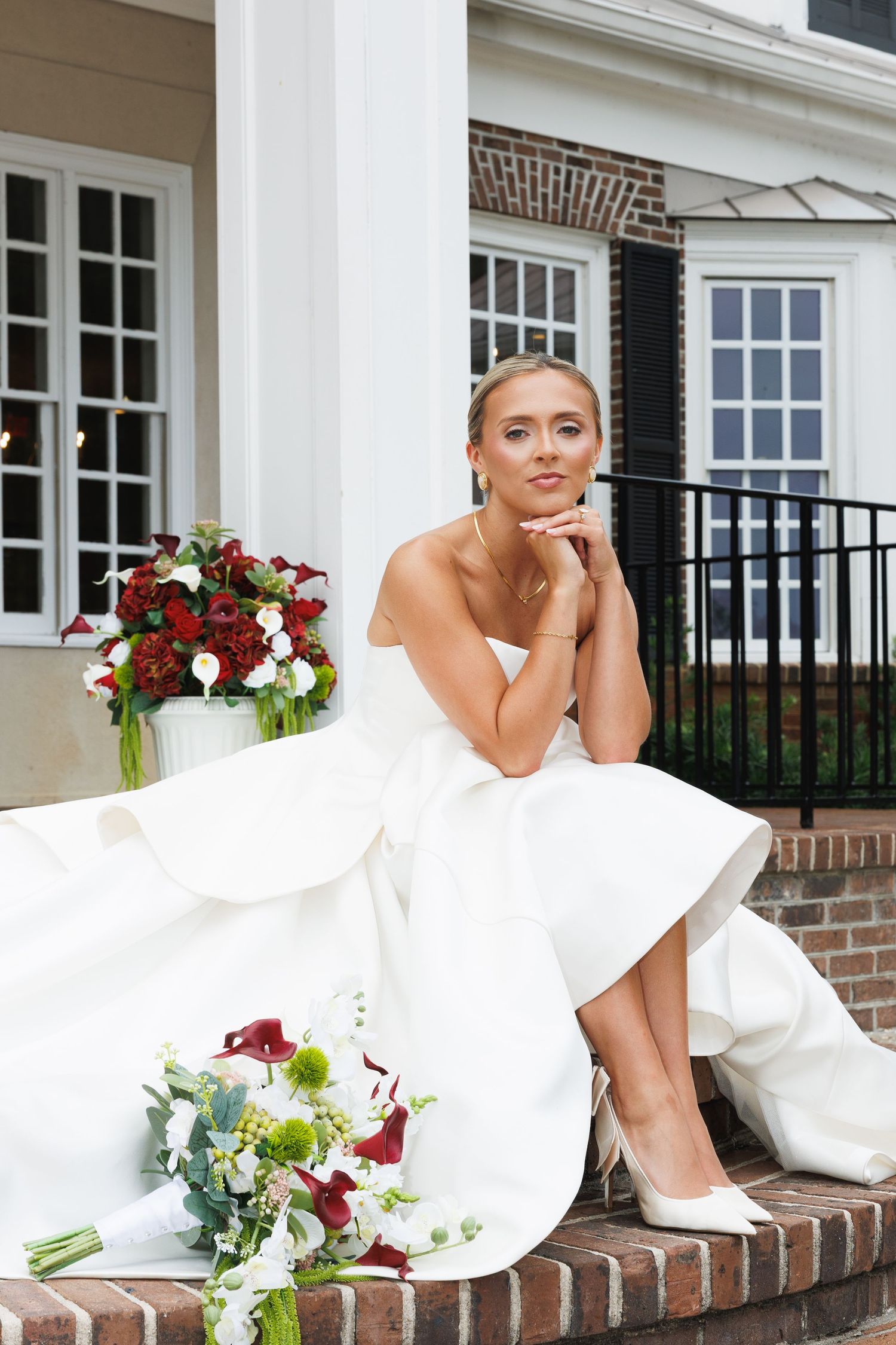 Bride in white strapless gown sits on brick steps with red and white bouquets outside elegant venue.