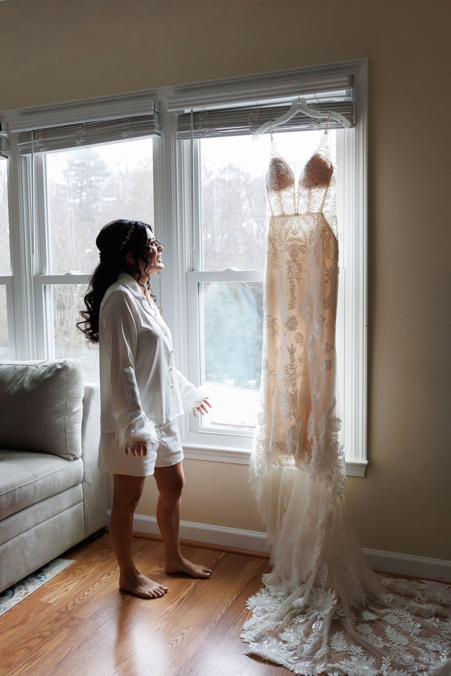 Bride in white robe admires her elegant lace wedding gown hanging by a bright window before ceremony.