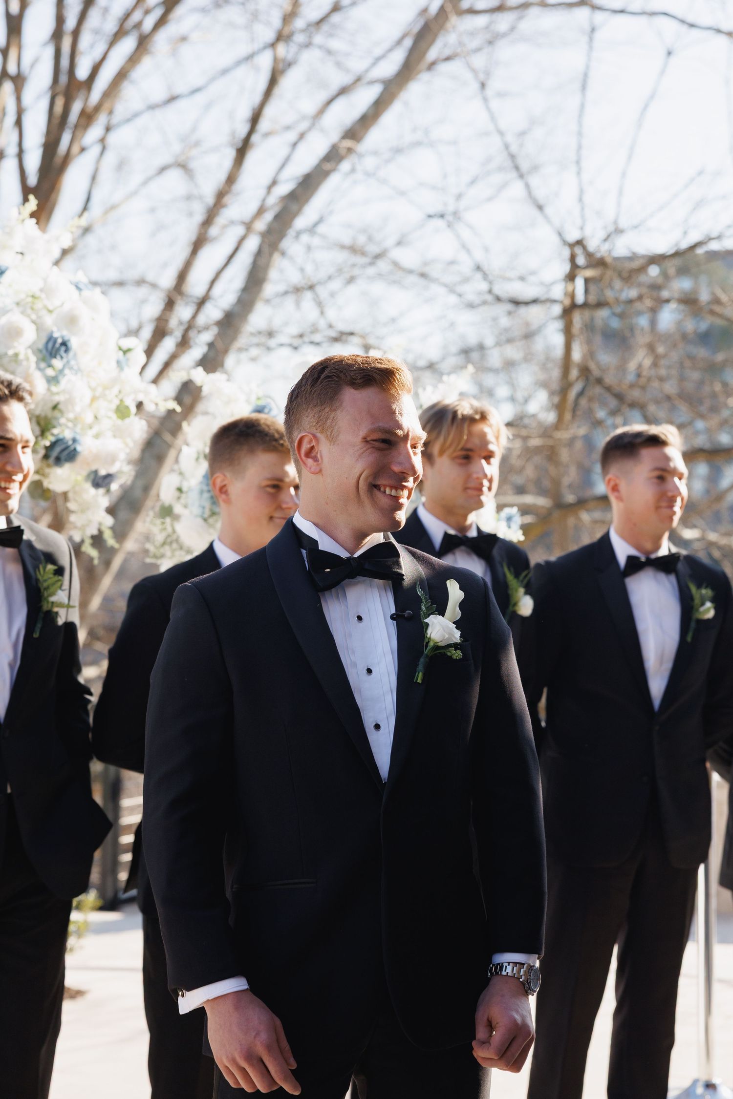 Smiling groom in black tuxedo with bow tie standing with groomsmen outdoors before ceremony.