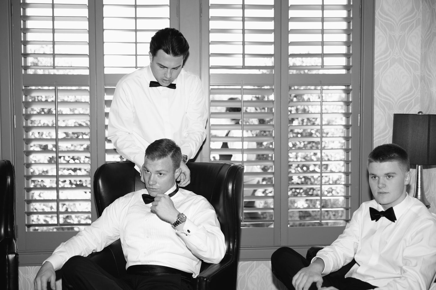 Three groomsmen in tuxedos prepare for a wedding in a black and white portrait photo.