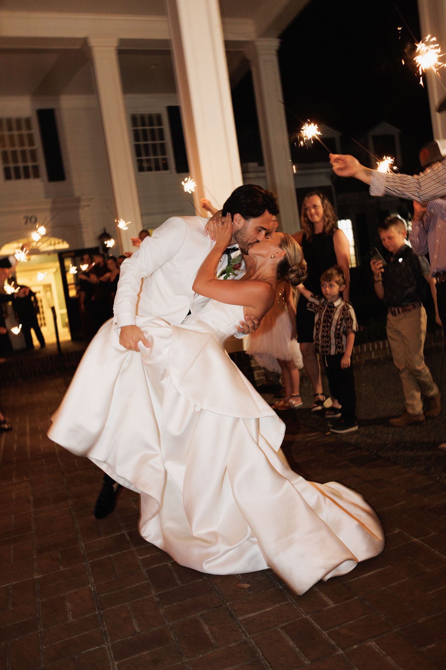 Groom dips bride for a romantic kiss during sparkler send-off outside a white columned venue at night.