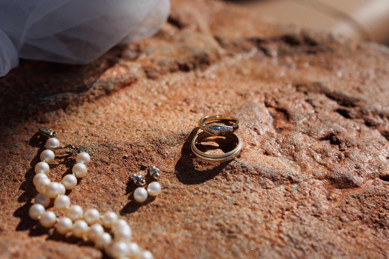 Gold wedding rings and pearl necklace with earrings displayed on rustic red sandstone surface.