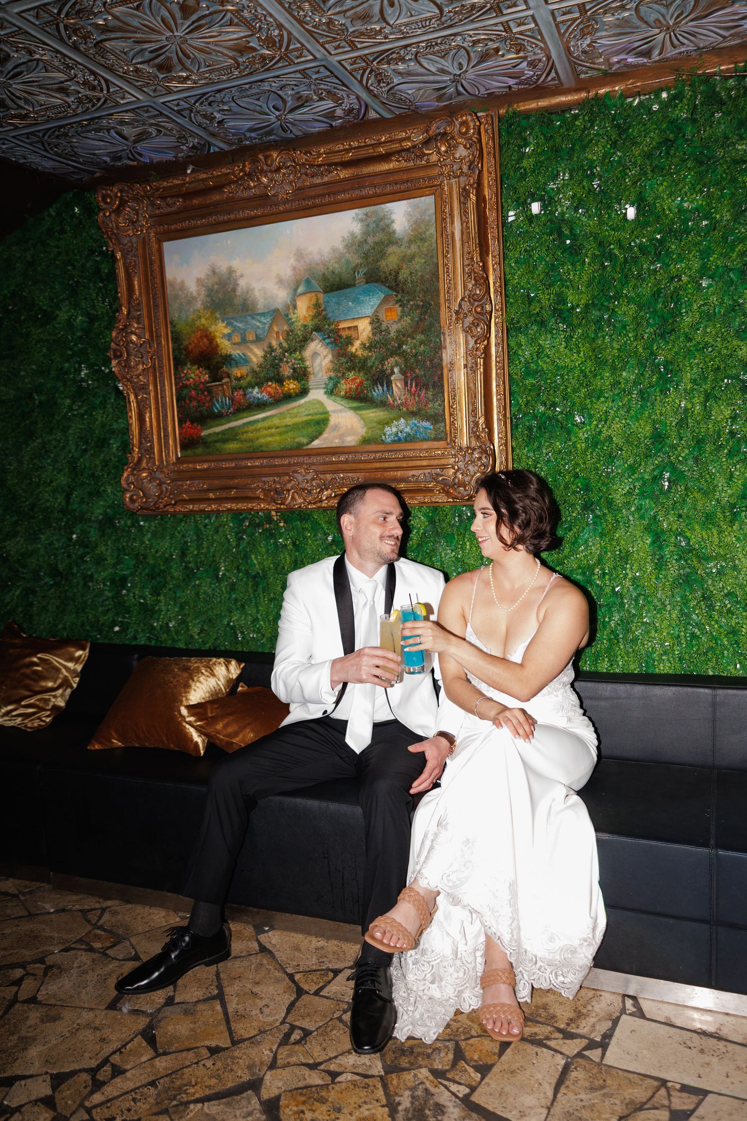 Bride and groom sitting together on a black sofa in front of a green moss wall with an ornate painting.