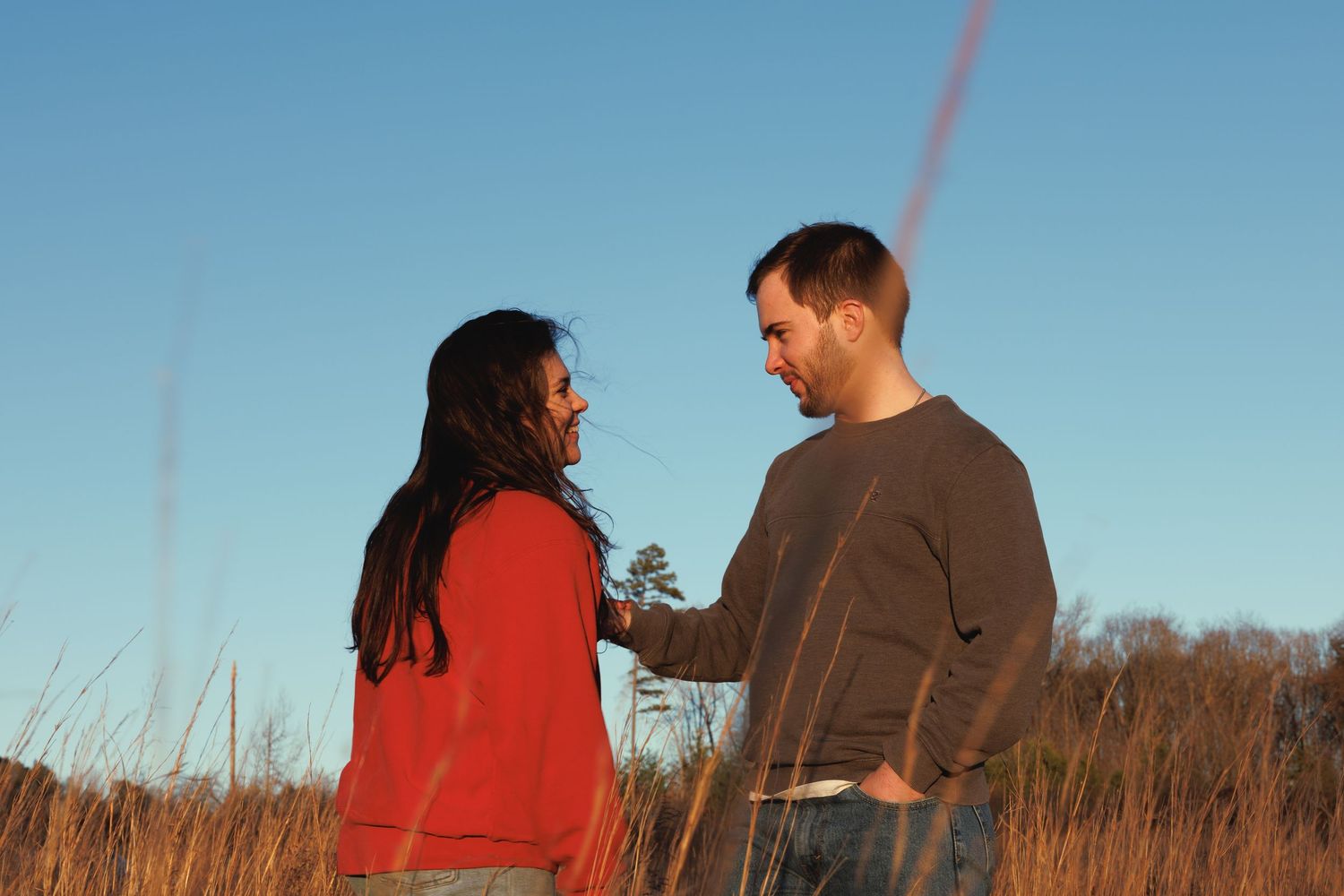 A couple stands facing each other in a golden field under a clear blue sky, sharing a tender moment.
