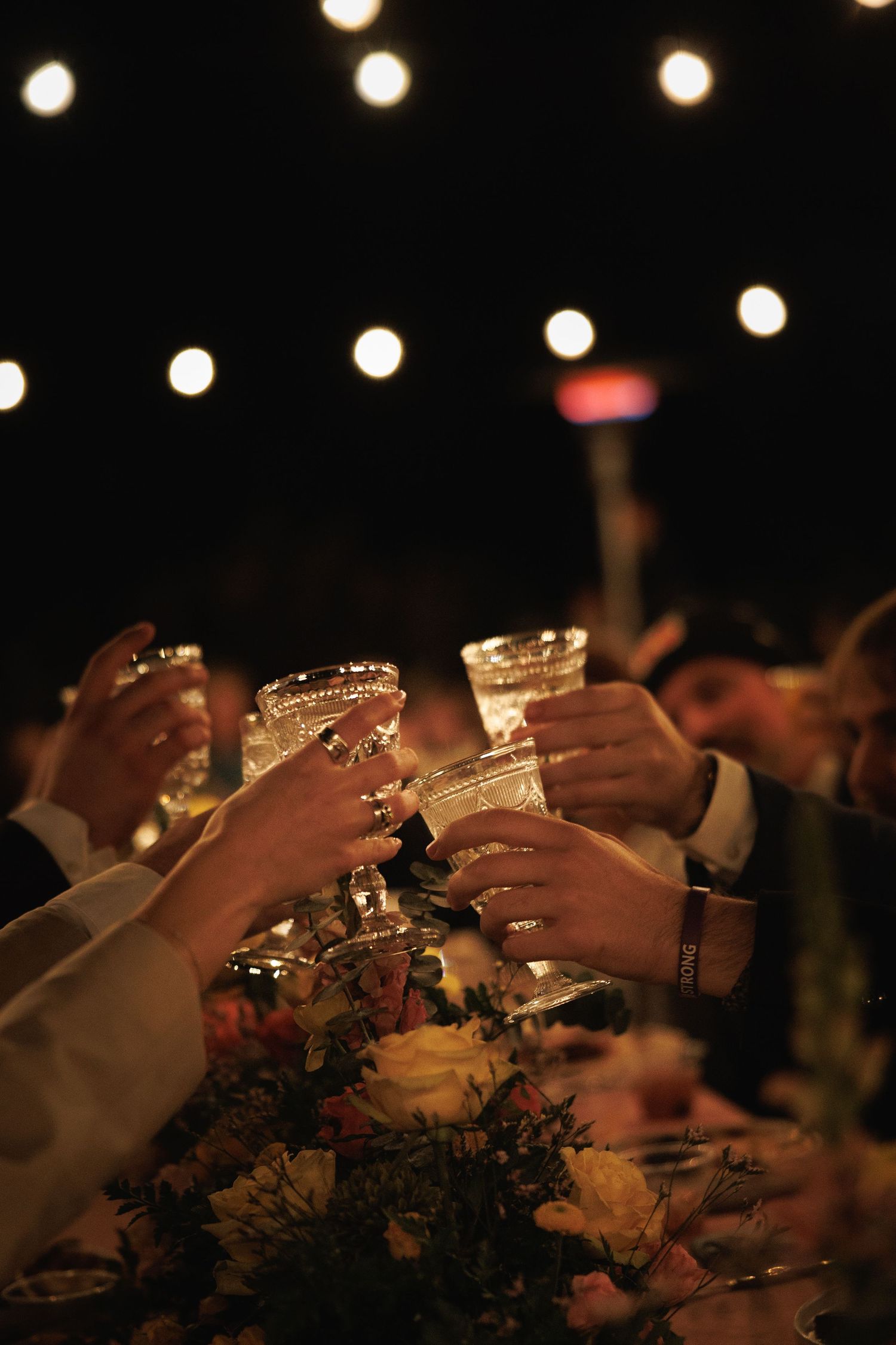 Wedding guests raising crystal glasses in toast under warm string lights at outdoor reception.