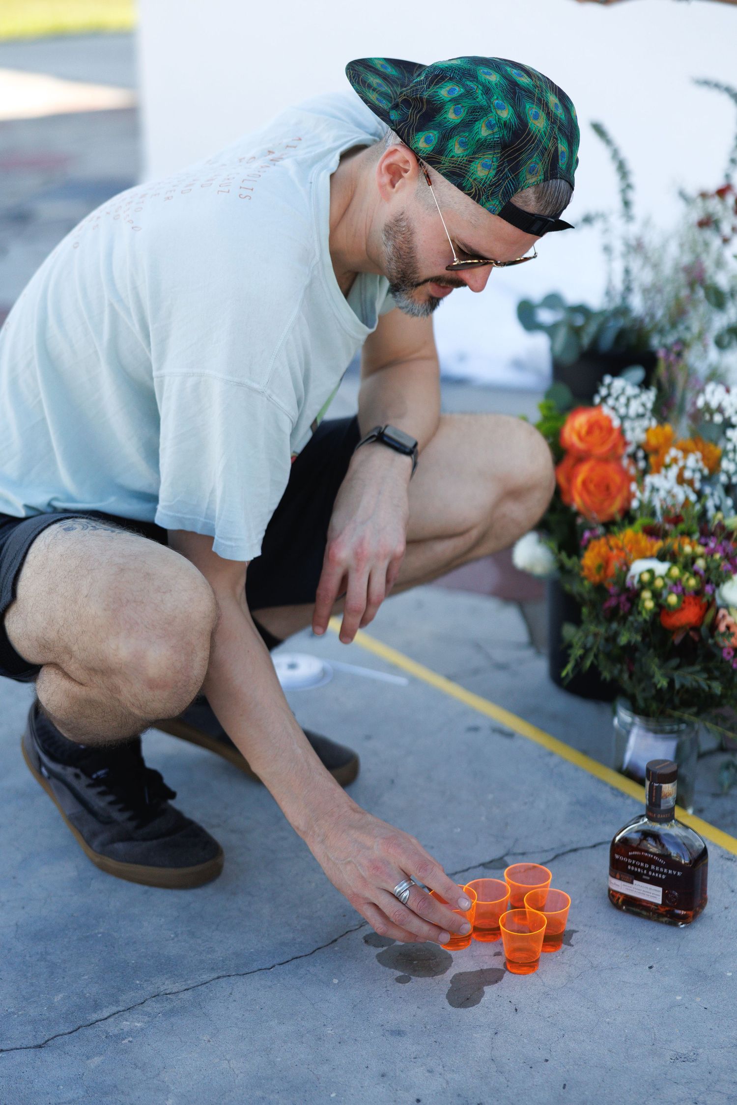 Man crouching to place orange cups and whiskey bottle at an outdoor memorial with flowers.