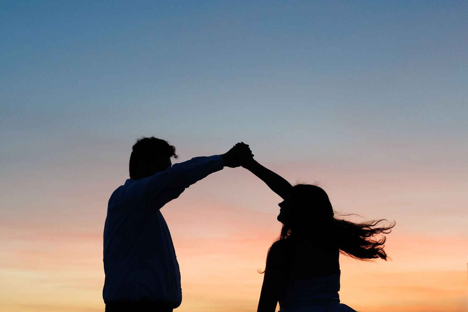 Romantic couple silhouette dancing at sunset, holding hands against colorful twilight sky.