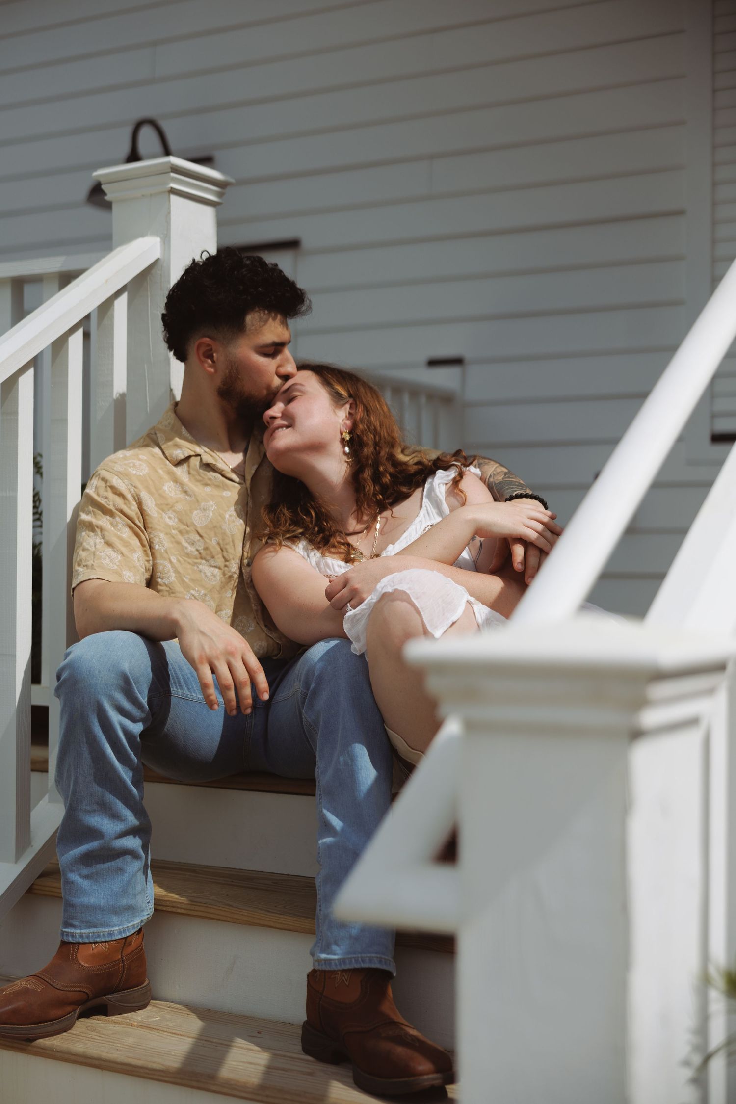 Romantic couple sharing a kiss on white porch steps, man in floral shirt and jeans, woman in white dress, sunny day.