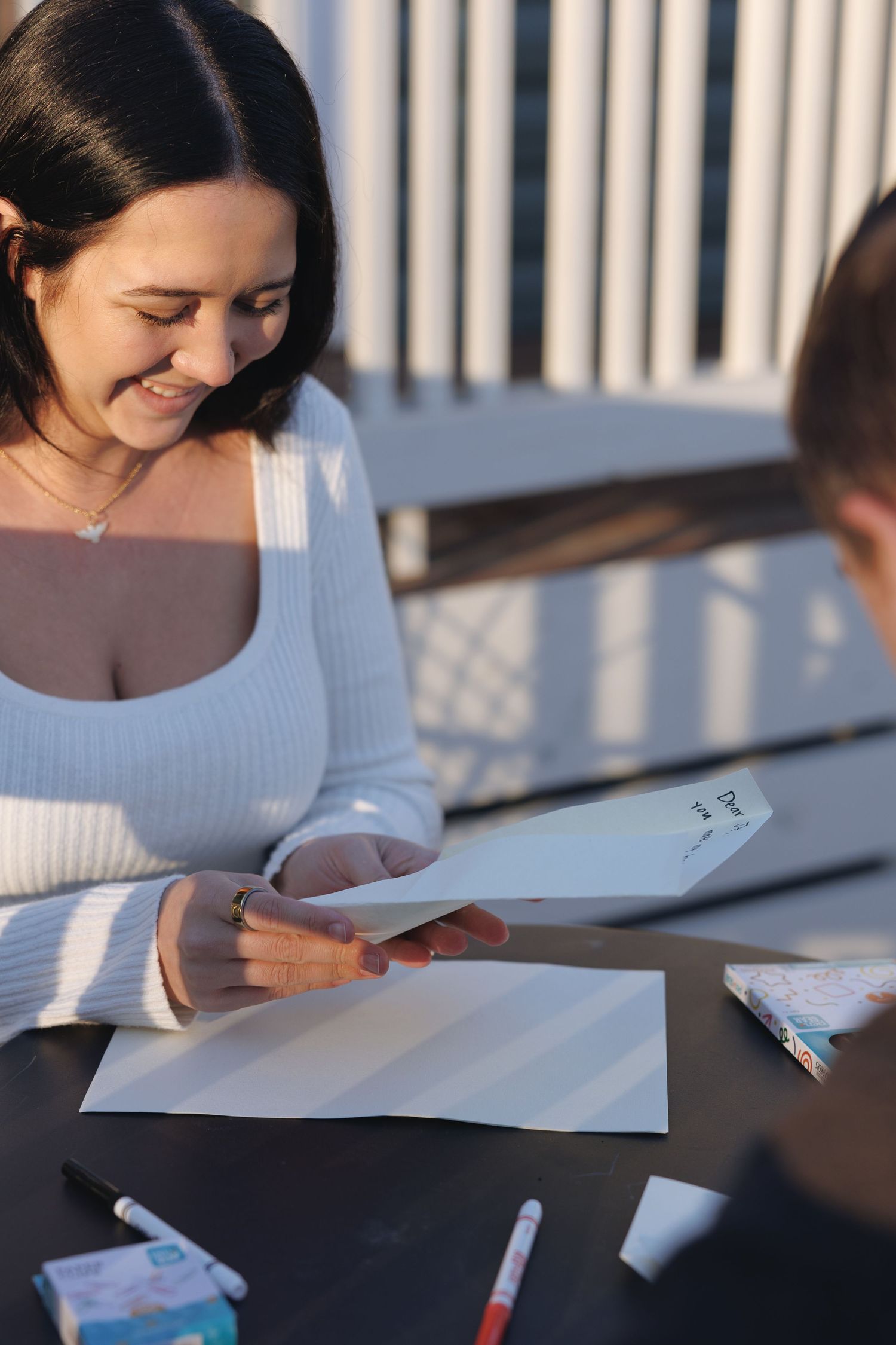 Smiling woman in white sweater reading a card at outdoor table with markers in warm sunlight.