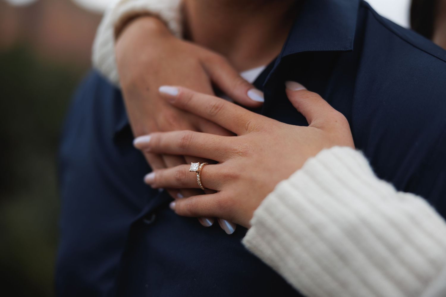 Close-up of woman's hand with gold engagement ring resting on man's chest in blue shirt.