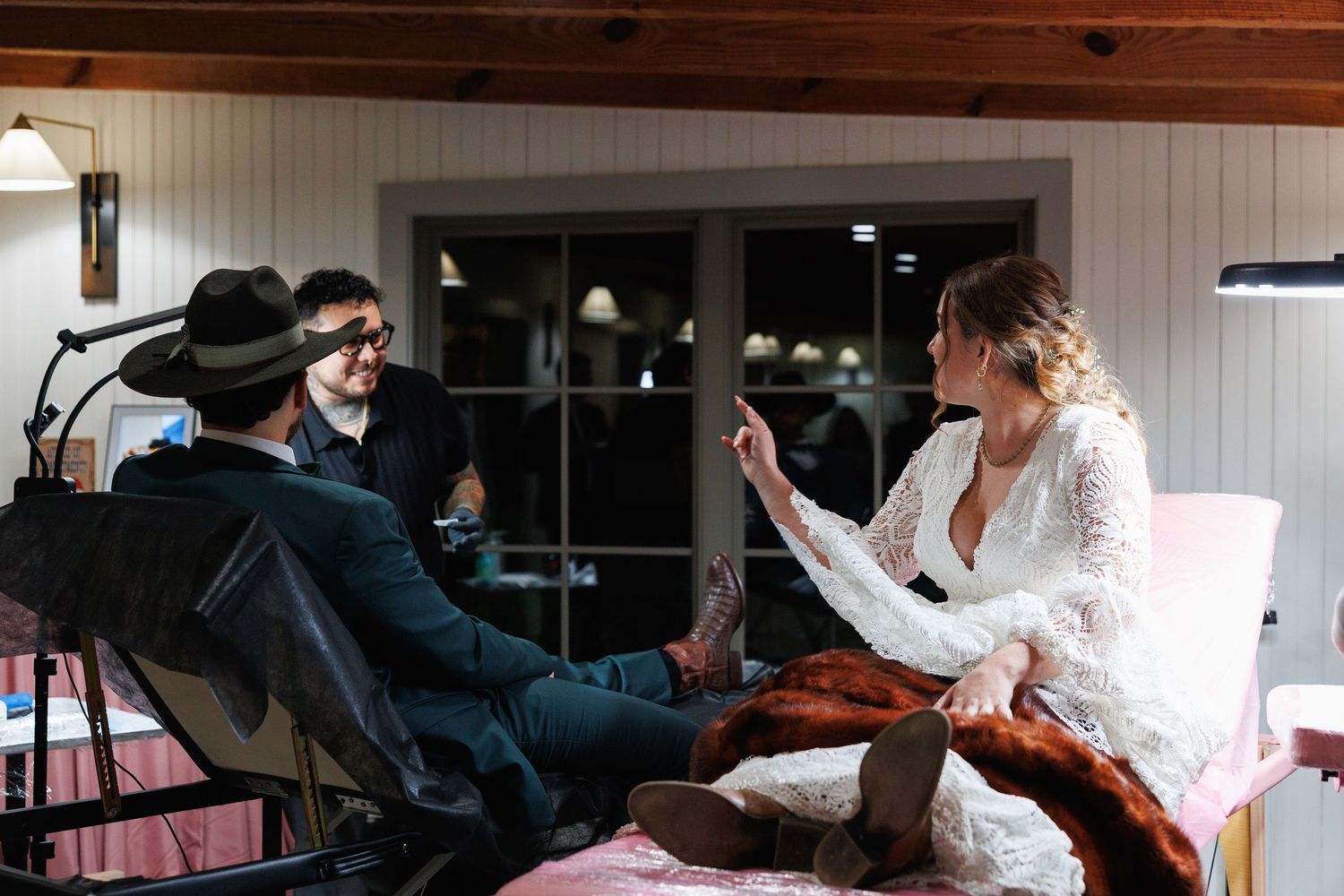 Bride in lace wedding gown sits with groom in hat during relaxed pre-wedding preparations indoors.