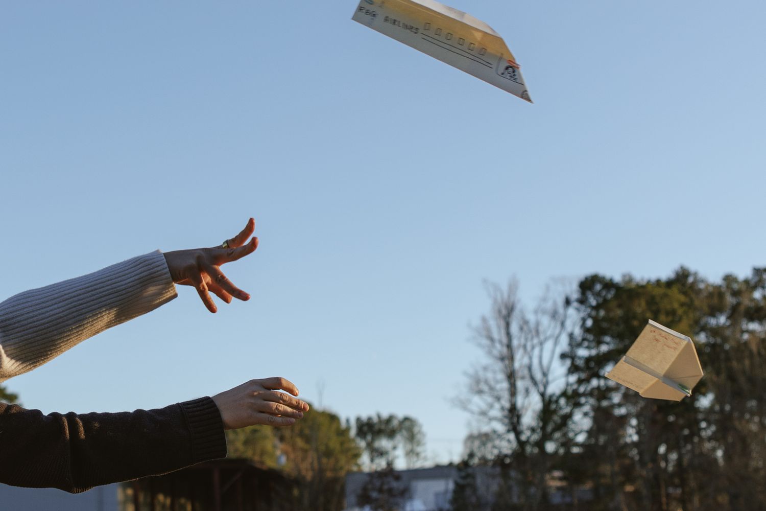 Two hands tossing paper airplanes into a clear blue sky outdoors near trees.