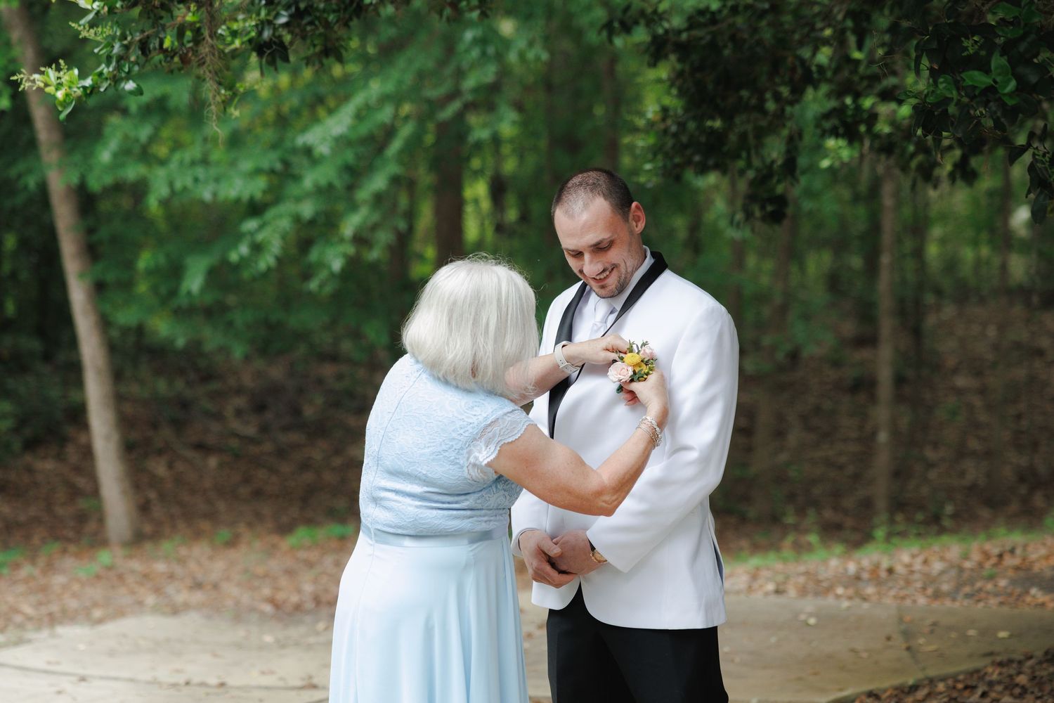 Groom in white tuxedo jacket receives boutonniere from older woman in blue dress outdoors at wedding.