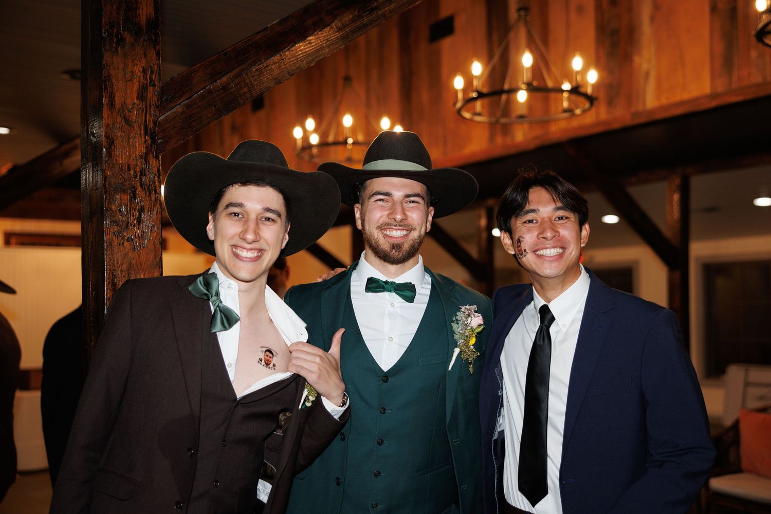 Three young men in suits and hats smiling together at rustic indoor wedding reception venue.
