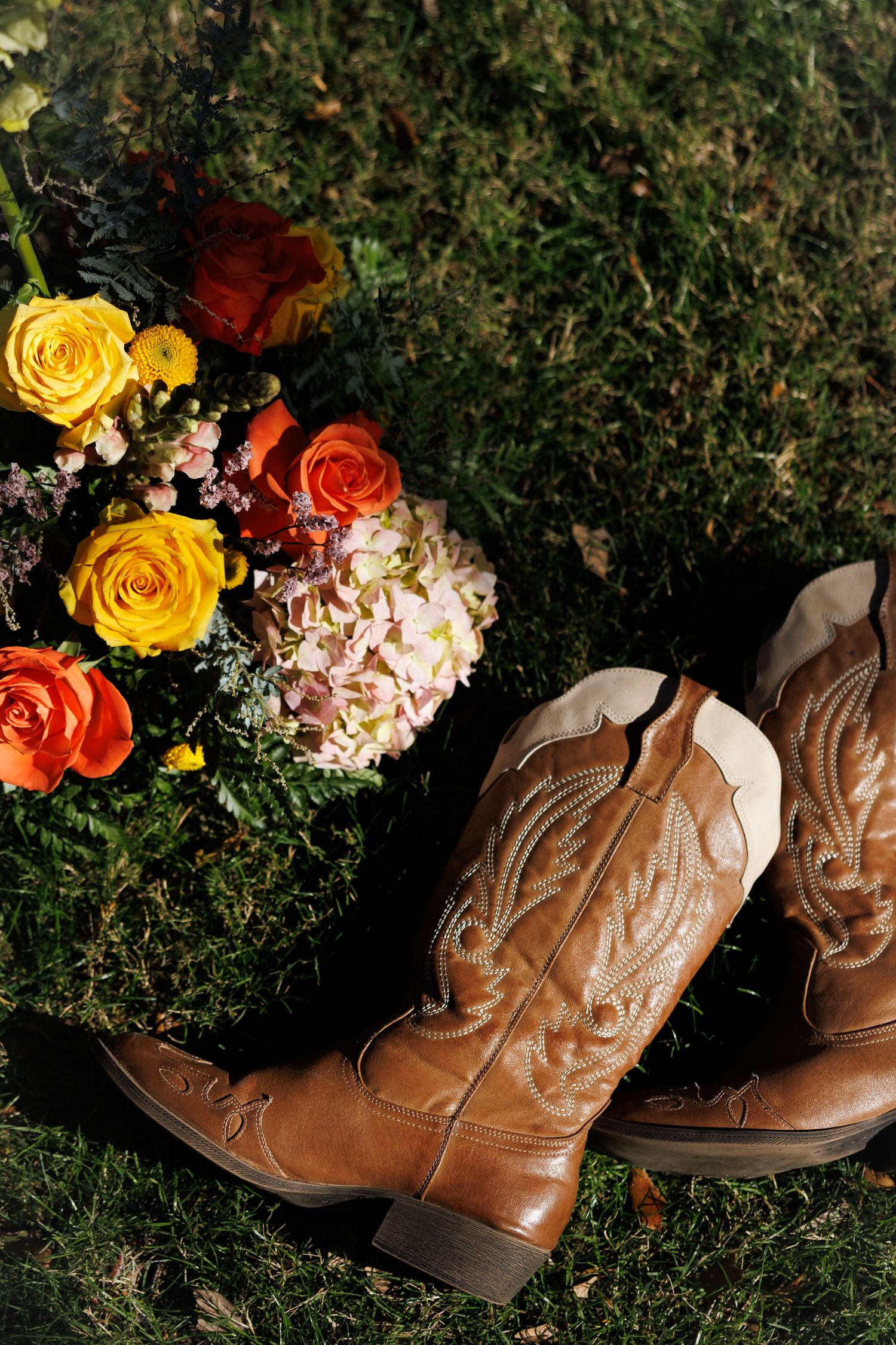 Brown cowboy boots beside a vibrant bouquet of orange, yellow roses and hydrangeas on green grass.