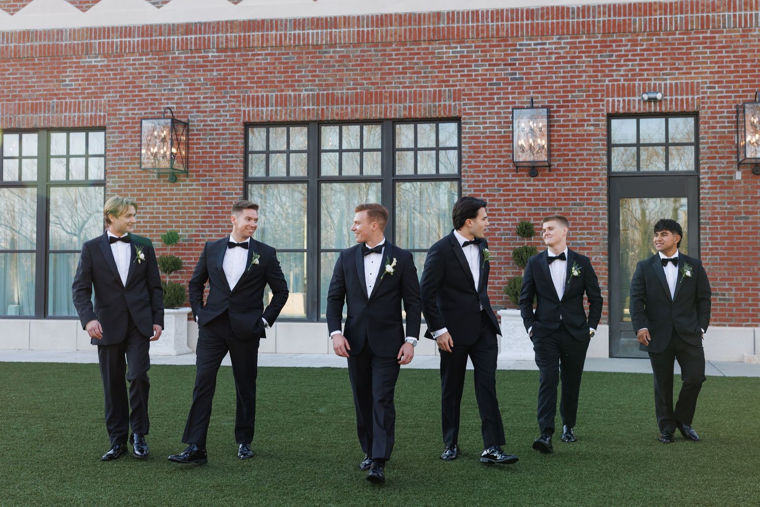 Groom and five groomsmen in matching black tuxedos pose casually outdoors in front of a brick building at The Yorkmont Courtyard in NC.