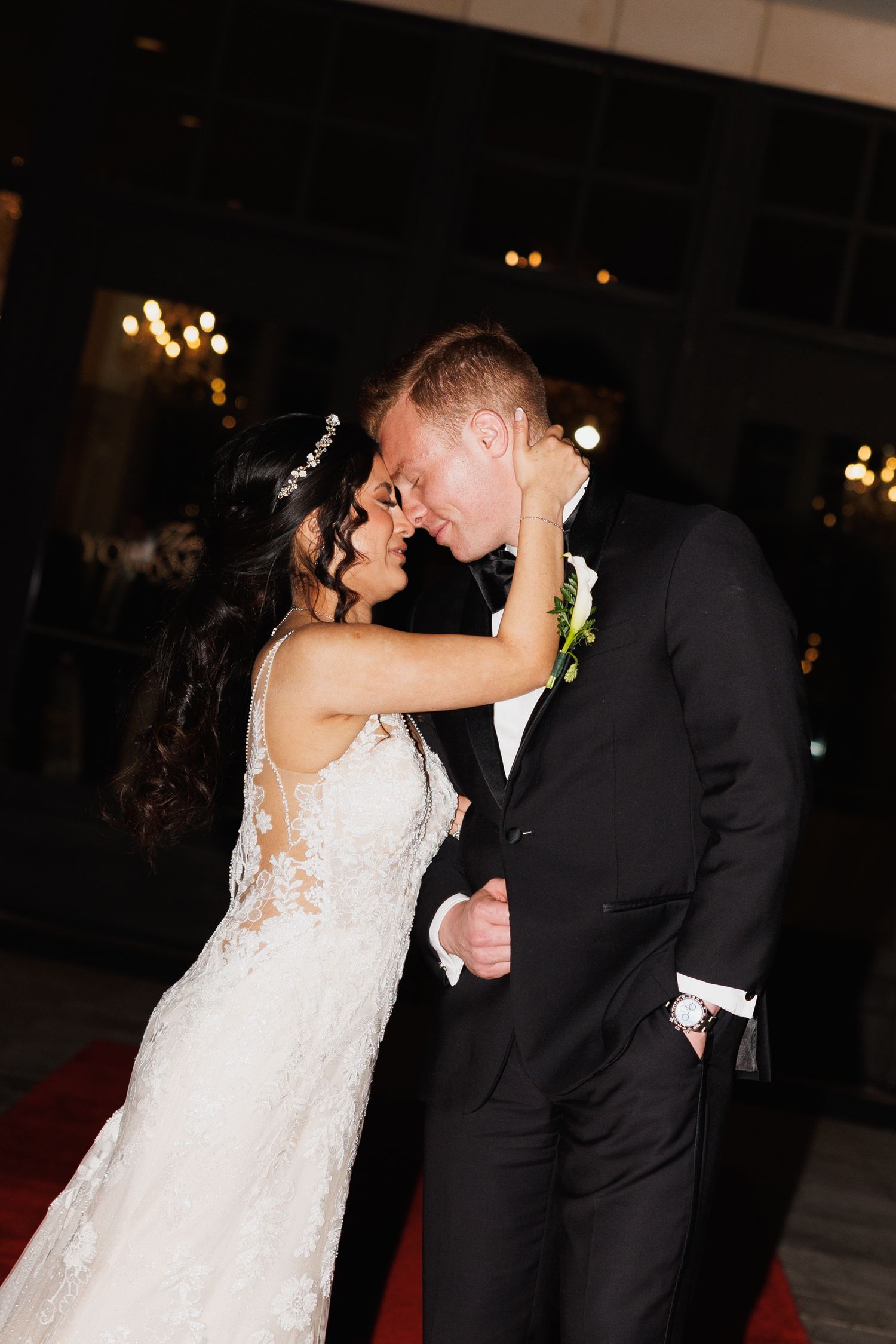 Bride in lace gown and groom in black tuxedo share romantic moment, nearly kissing on red carpet at night at The Yorkmont in Charlotte, NC.