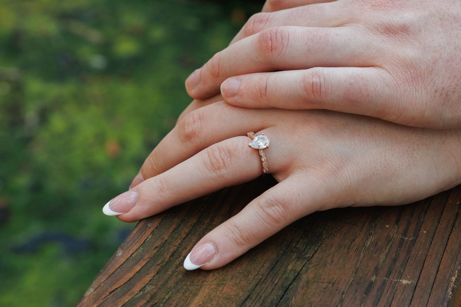 Rose gold oval diamond engagement ring on a woman's finger resting on a wooden surface outdoors.